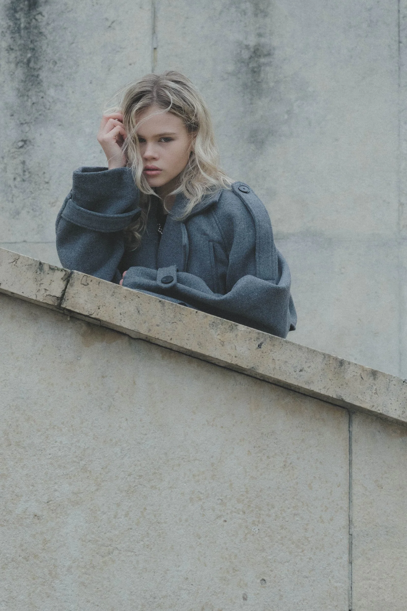Jeune femme aux cheveux blonds, portant un manteau gris, posant près d'un mur en béton.