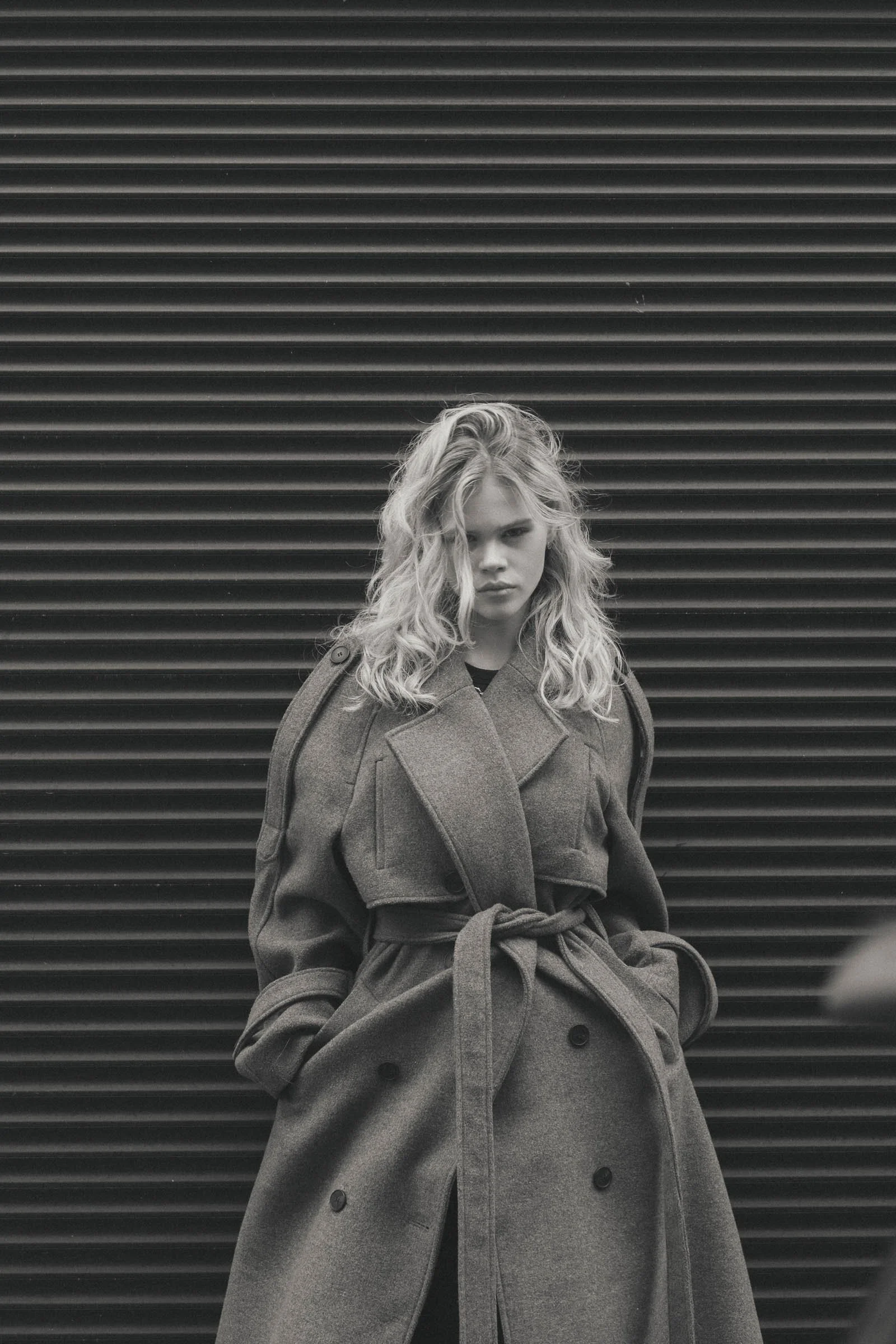 Une femme en manteau long en laine, lien à la taille, devant une façade en volets en métal, photo en noir et blanc.