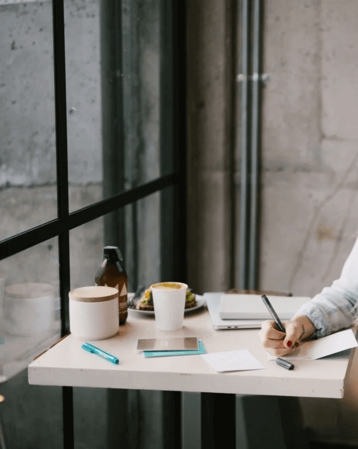 A modern workspace with a white table, a Squarespace website designer and health and wellness copywriter writing in a notebook, with a coffee cup, a closed laptop, a book, a blue pen, set in front of a large window with a concrete wall.