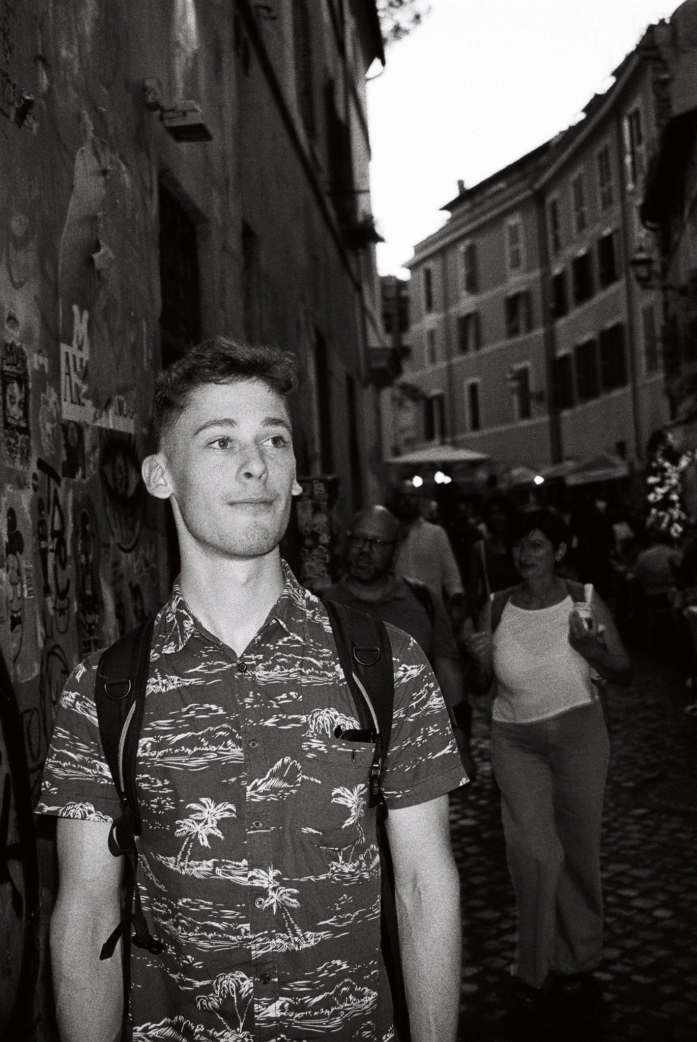 Black and white photo of a person with a backpack standing on a narrow, cobblestone street lined with old buildings. The person is wearing a patterned shirt and gazing upwards. In the background, several people are walking, and the walls are covered 