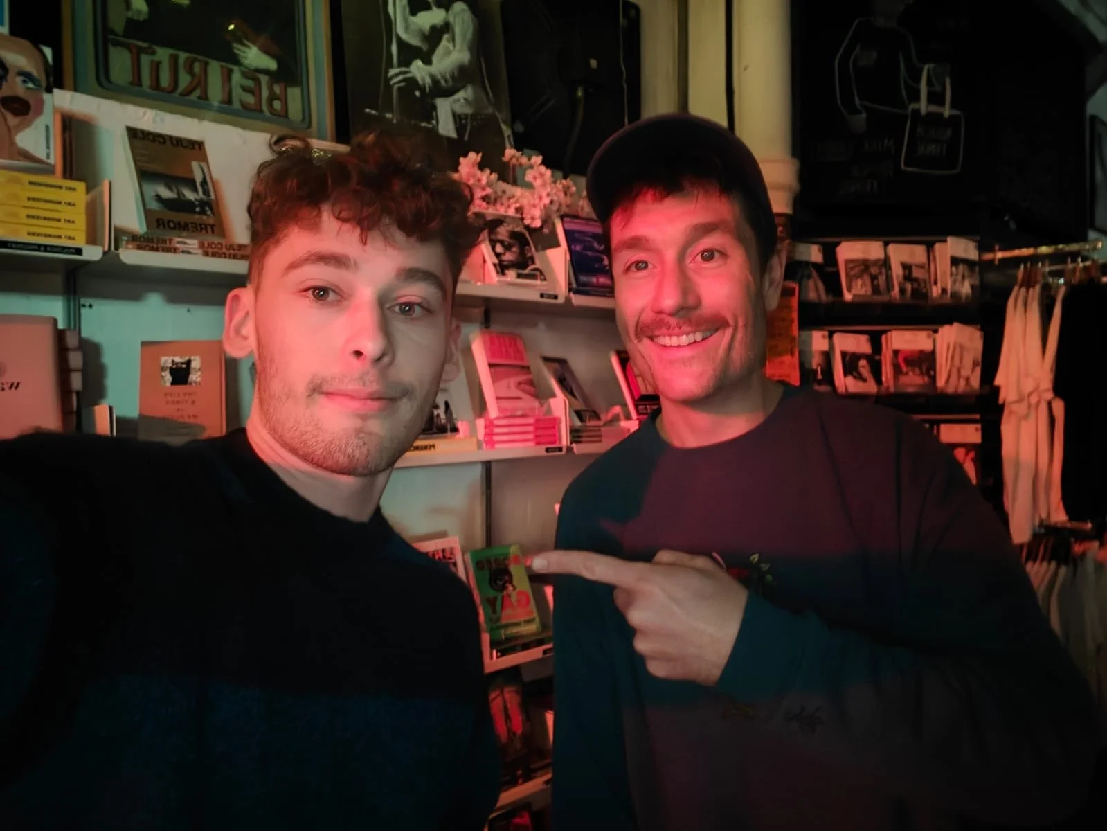 Two people posing in a bookstore with bookshelves in the background, one person pointing at the other.