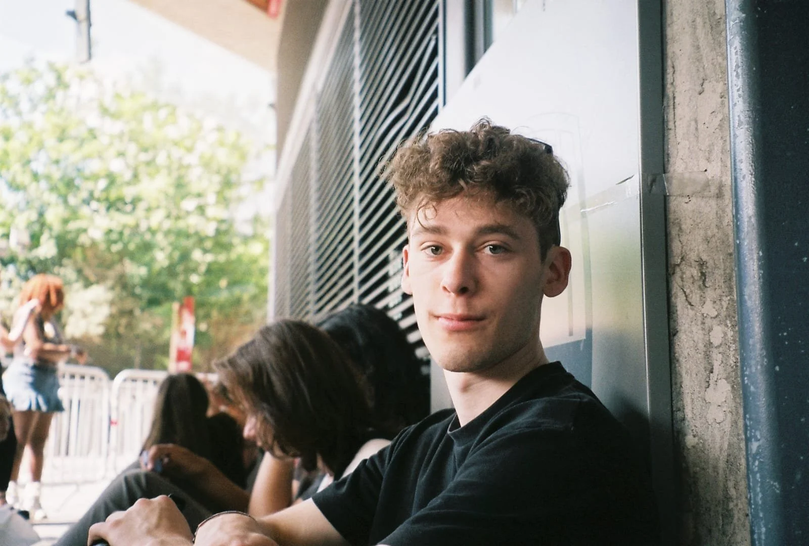Young man with curly hair sitting outdoors, leaning against a wall, with people in the background.