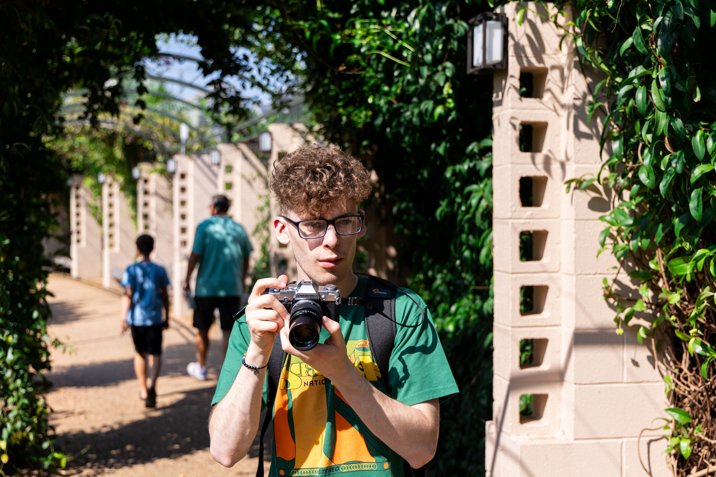 Person holding camera in a garden setting with people walking in the background.