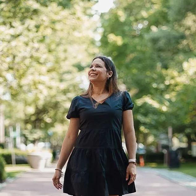 Woman in black dress walking outdoors, smiling, surrounded by trees.