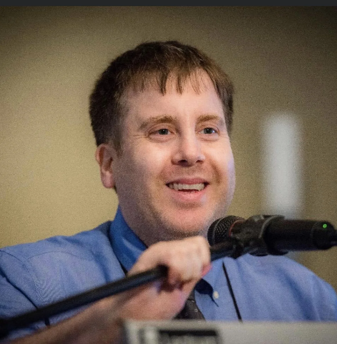 A man with brown hair and blue eyes, wearing a blue shirt, smiling while speaking into a microphone.