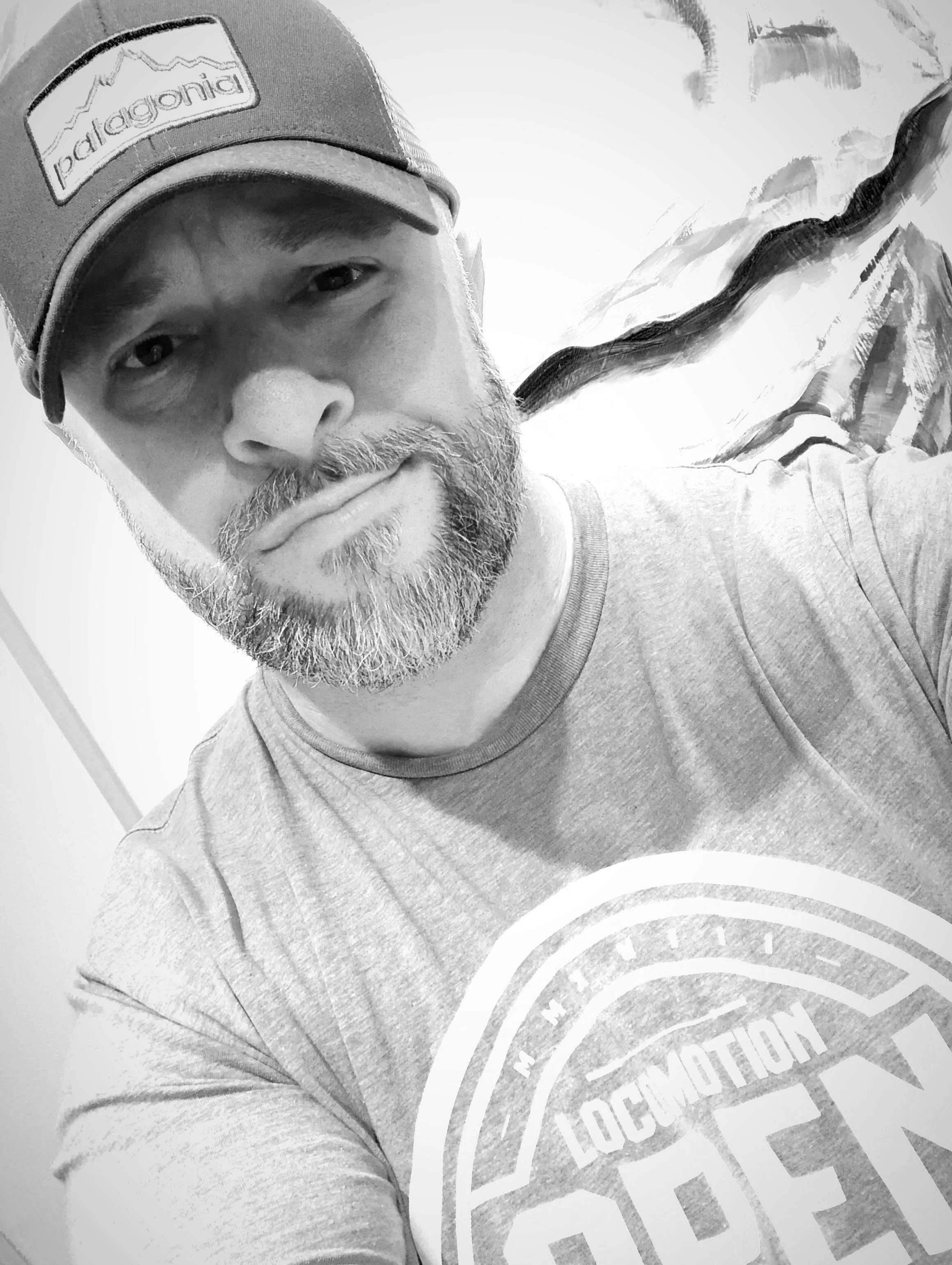 A black and white close-up selfie of a man wearing a baseball cap with a Patagonia logo and a t-shirt with a circular logo.