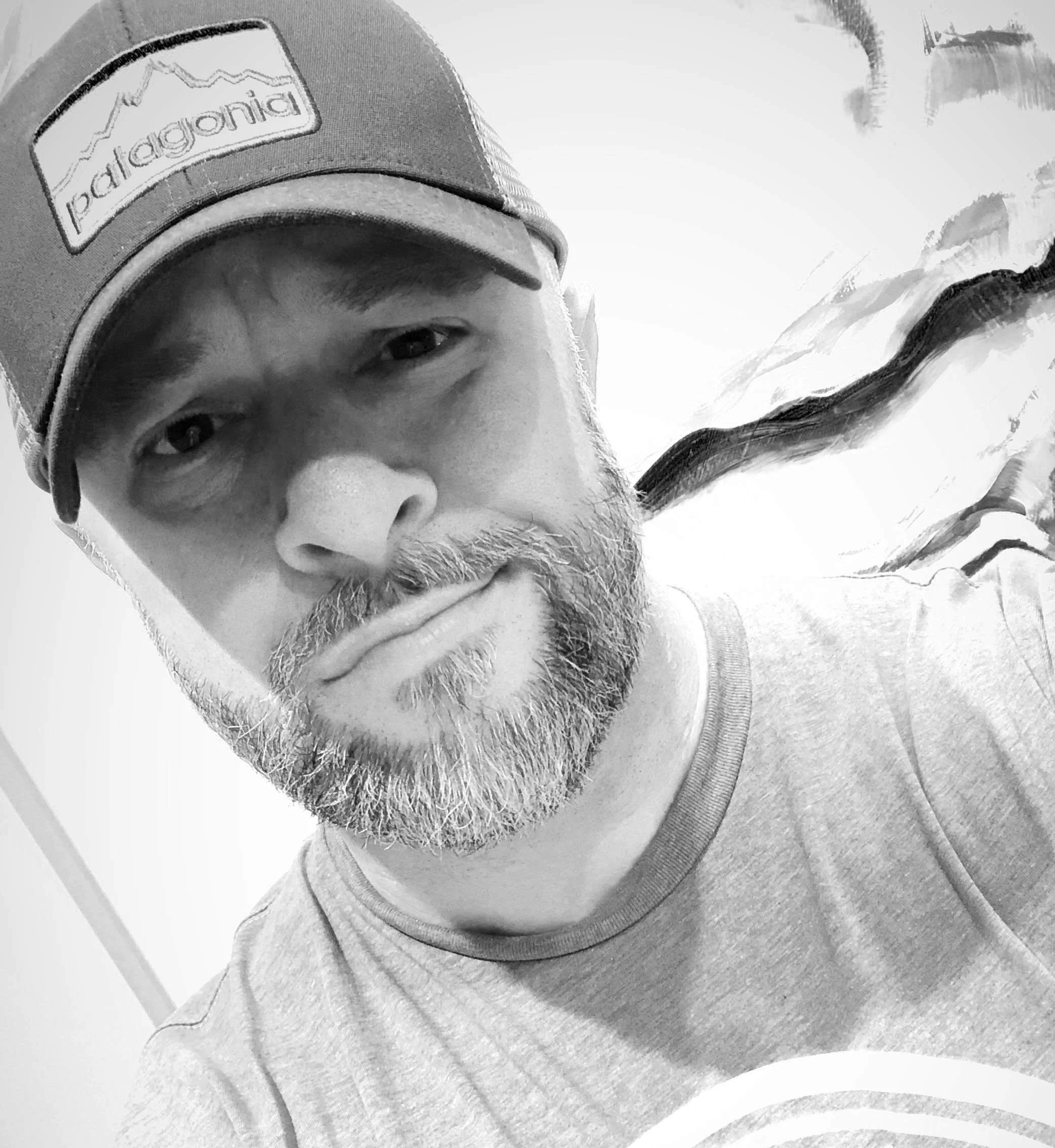 Black and white selfie of a man wearing a baseball cap with the Patagonia logo, with a beard, looking at the camera with a skeptical expression.