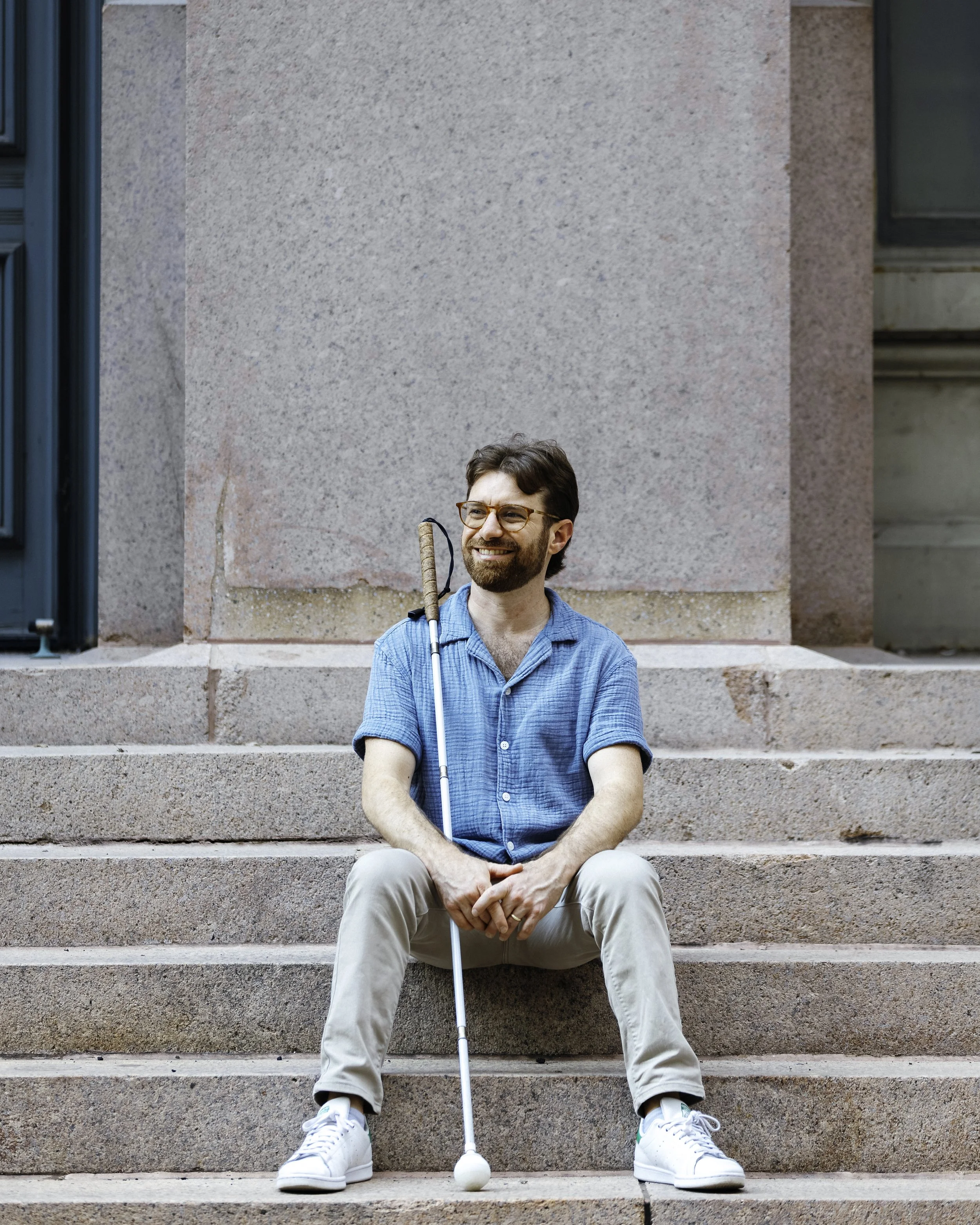 Charlie is sitting on stone steps and is looking into the distance. He is smiling and is holding his cane.