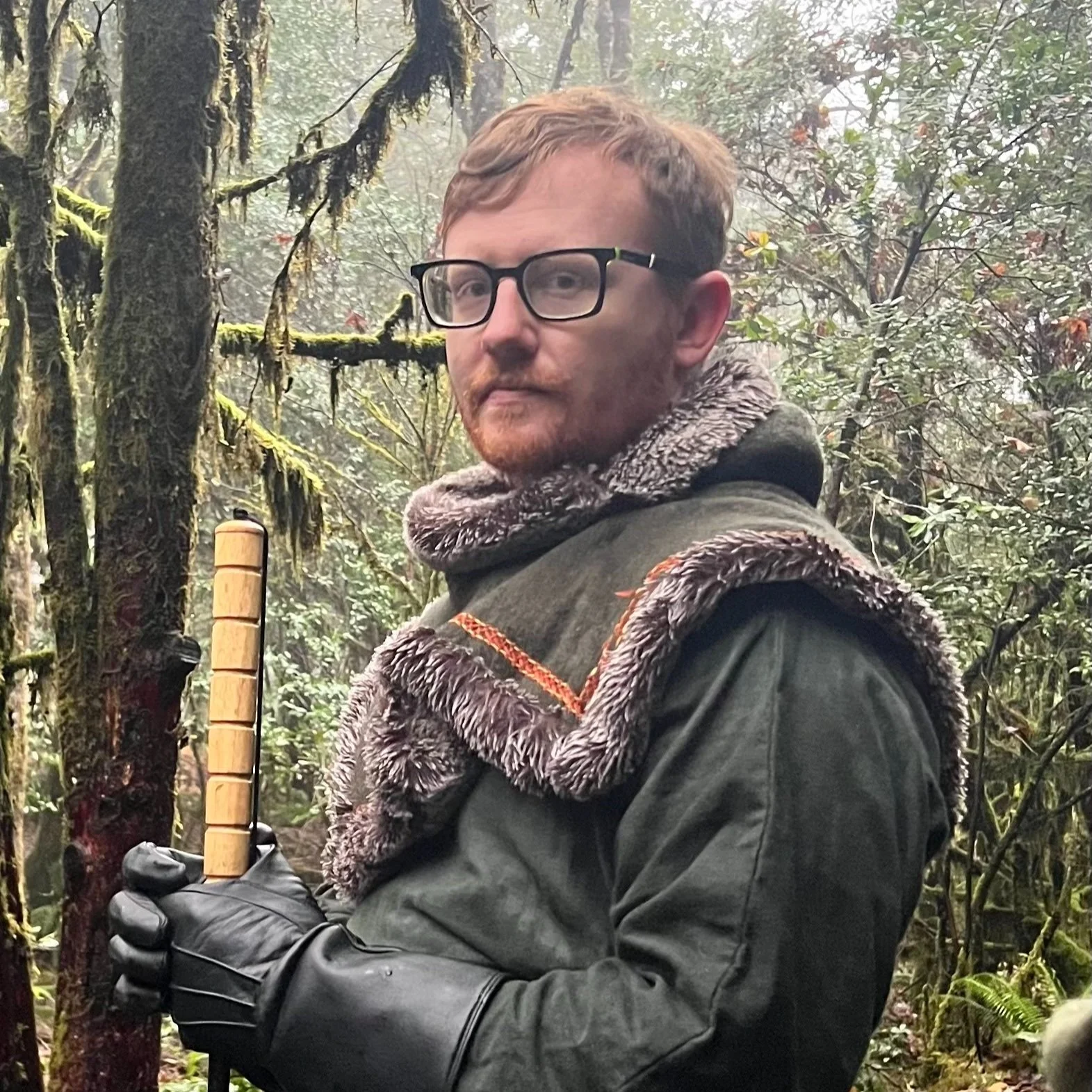 A person with glasses and a beard wearing a fur-lined jacket and black gloves standing in a mossy forest, holding a wooden-handled tool.