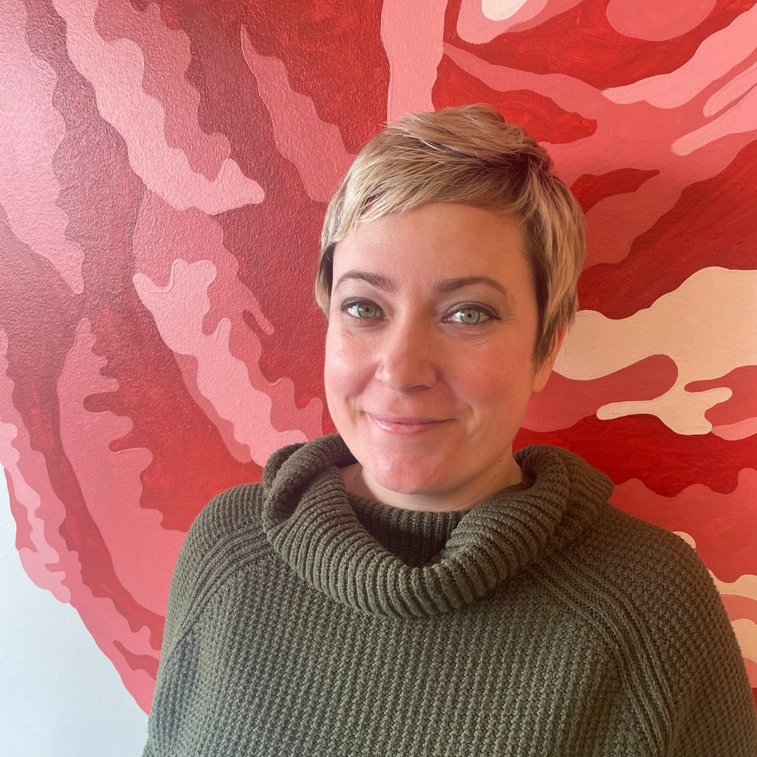 A woman with short blonde hair and green eyes, wearing a dark green ribbed turtleneck sweater, smiling in front of a red, pink, and white abstract patterned wall.