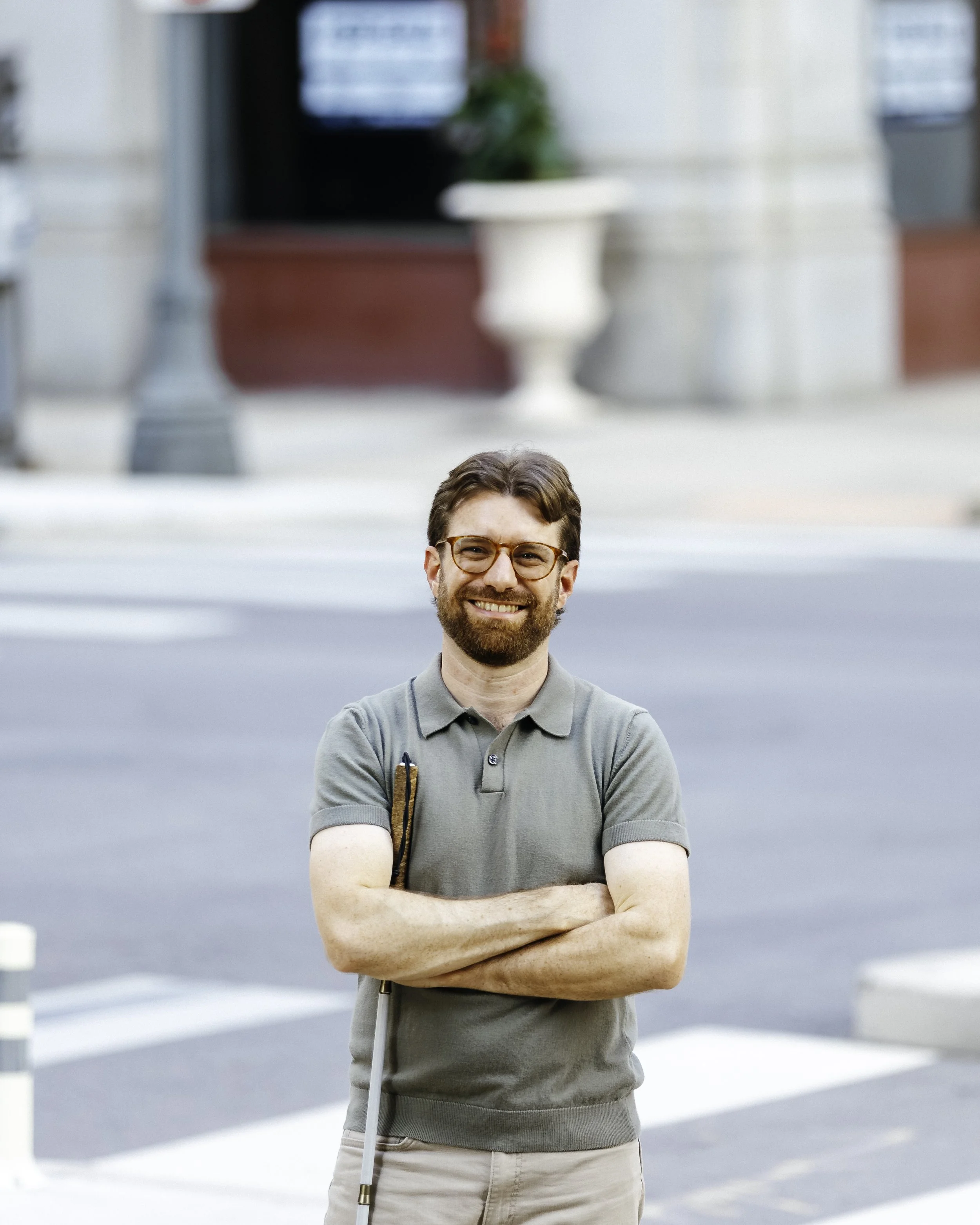 Charlie standing on the sidewalk in front of a crosswalk. He is smiling and is holding his cane while folding his arms.
