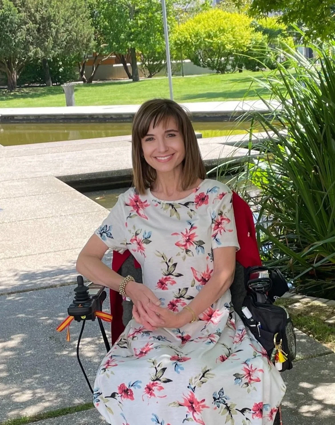 A woman with shoulder-length brown hair and a floral dress sitting on a mobility scooter outdoors near a pond and lush greenery.