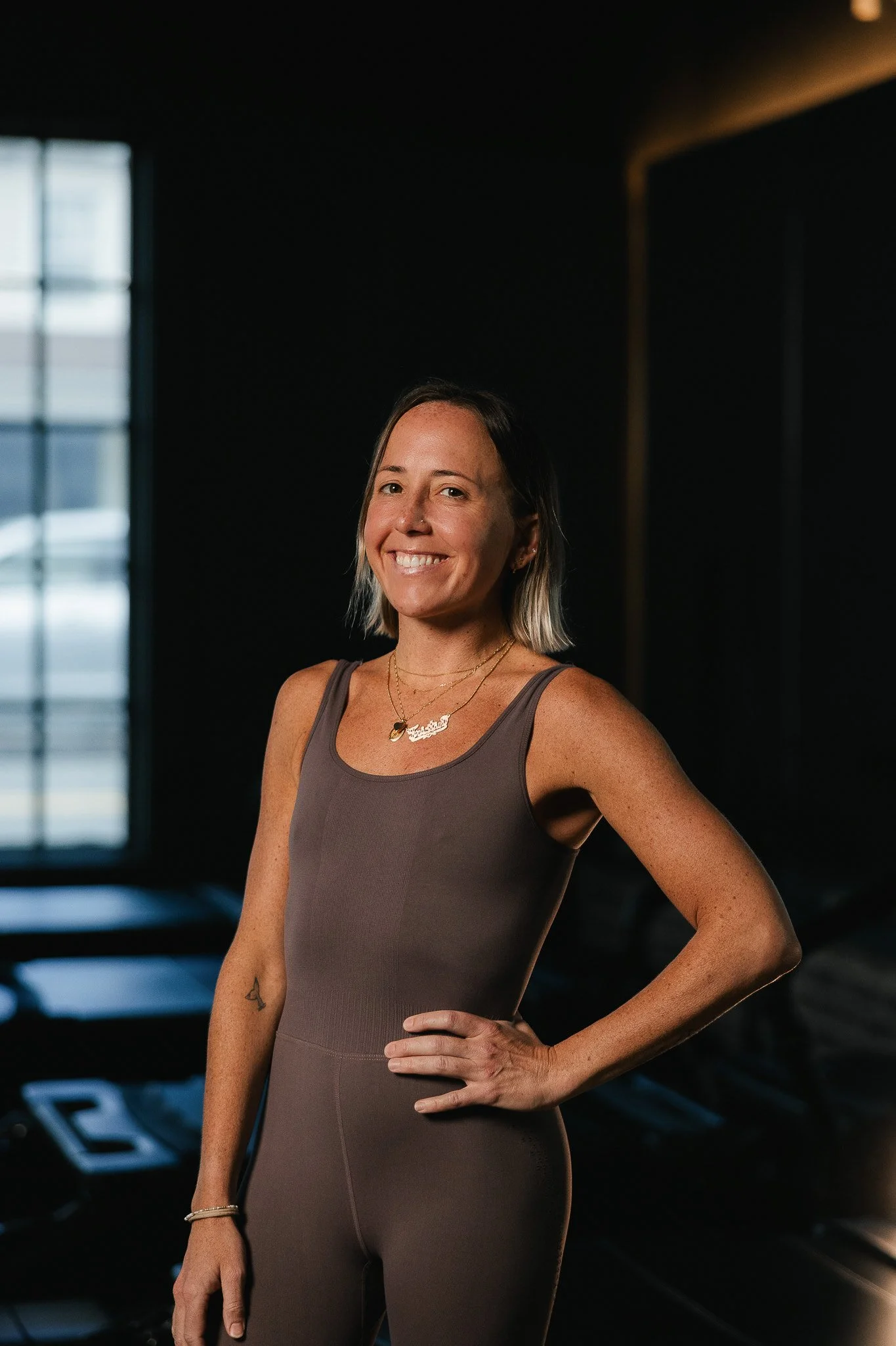 A woman in a brown sleeveless dress standing in a dark room, smiling, with her hand on her hip.