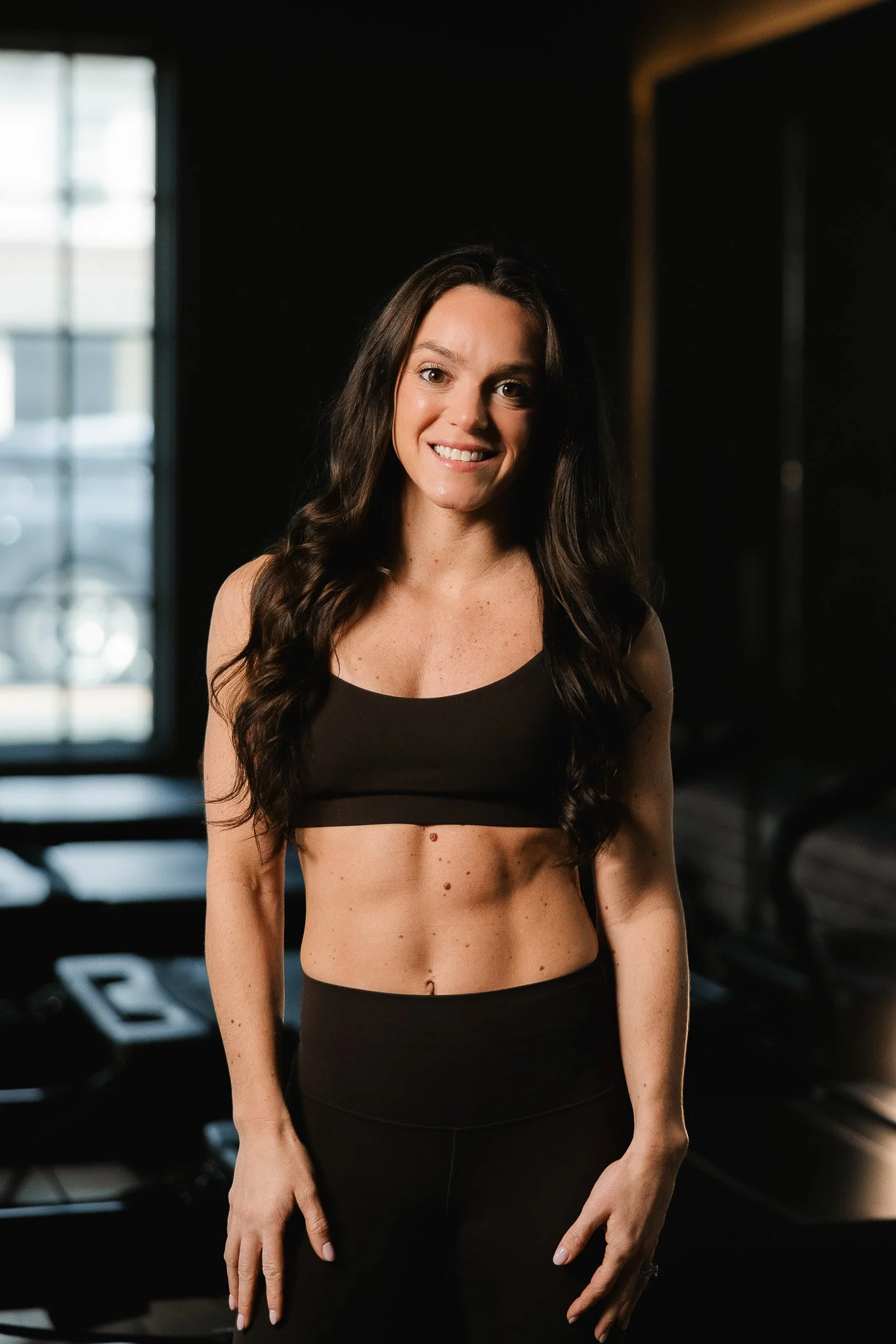 Woman with long dark hair in a gym, wearing a black sports bra and leggings, smiling at the camera.