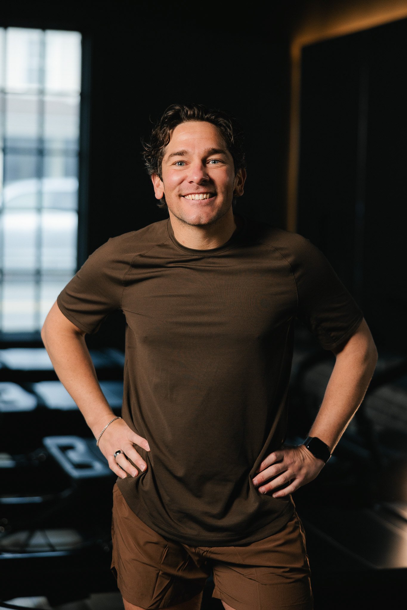 Smiling man with dark curly hair wearing a brown athletic t-shirt and brown shorts, standing inside a gym.