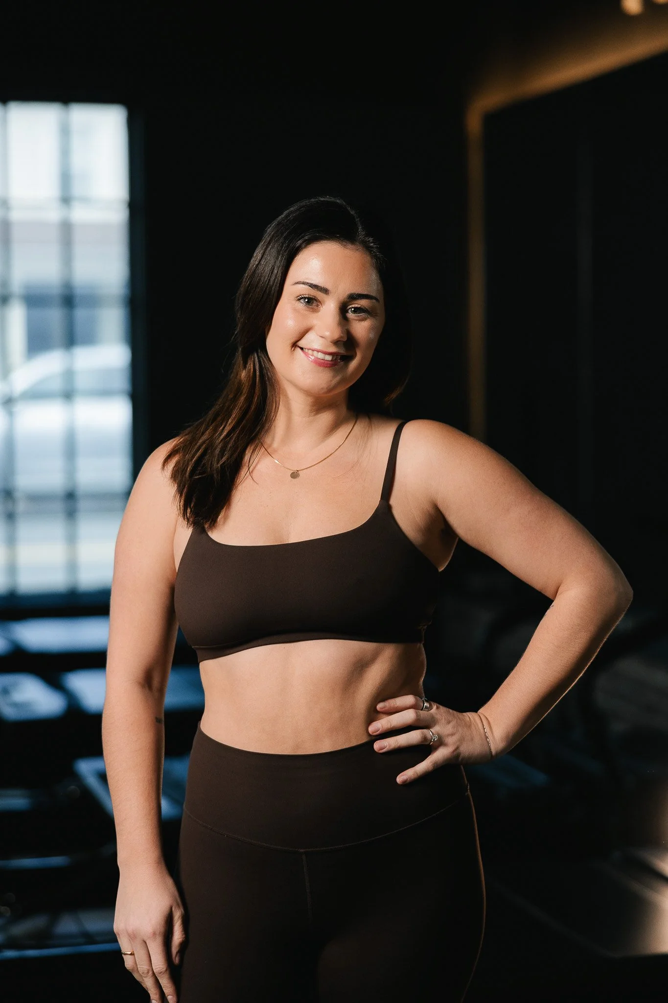 A woman with long dark hair wearing a black sports bra and black leggings, standing in a gym with equipment in the background, smiling with one hand on her hip.