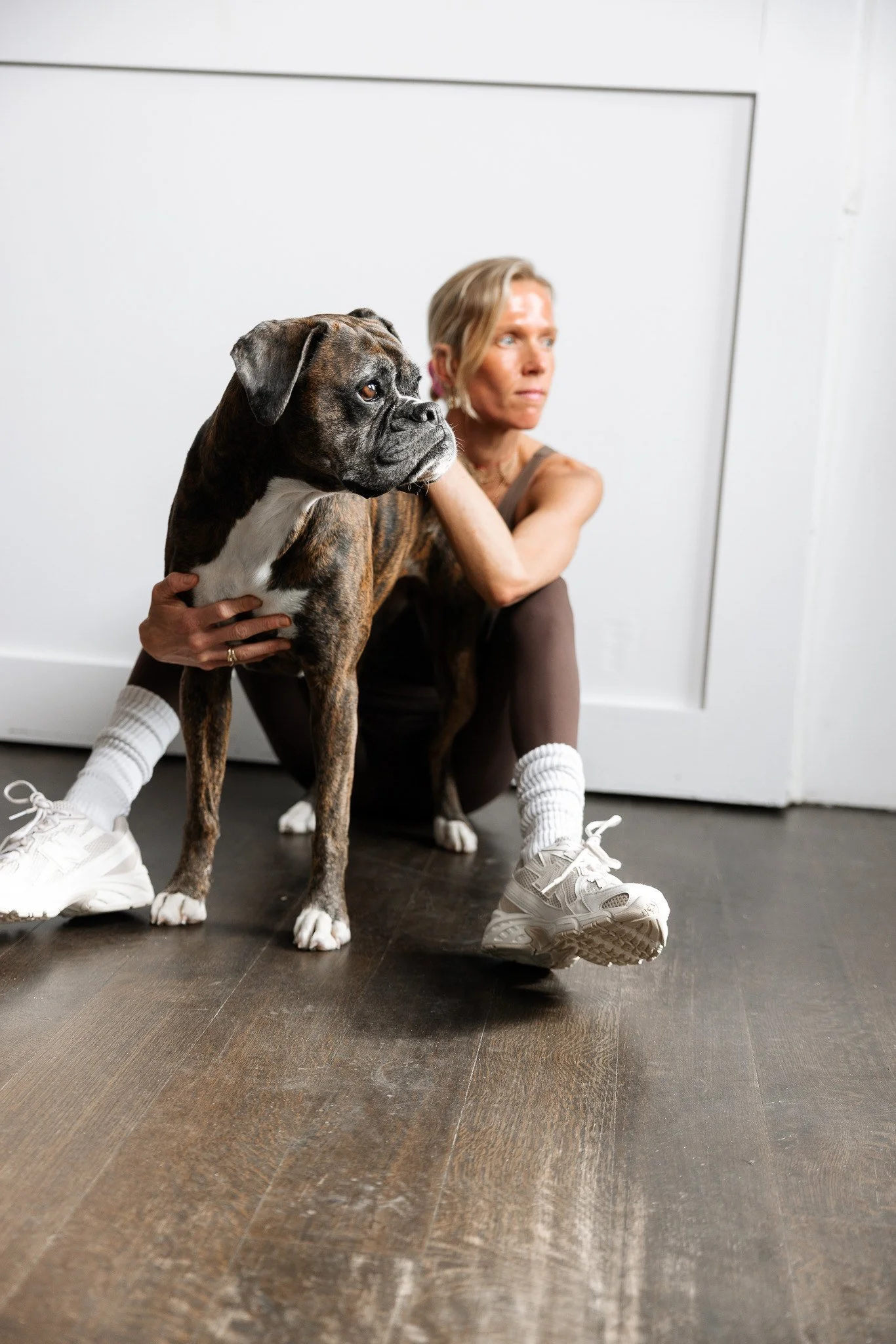 A woman in a sitting yoga pose with her chin resting on her knee, holding a large brindle Boxer dog close to her with one hand, on a dark wood floor against a white wall background.
