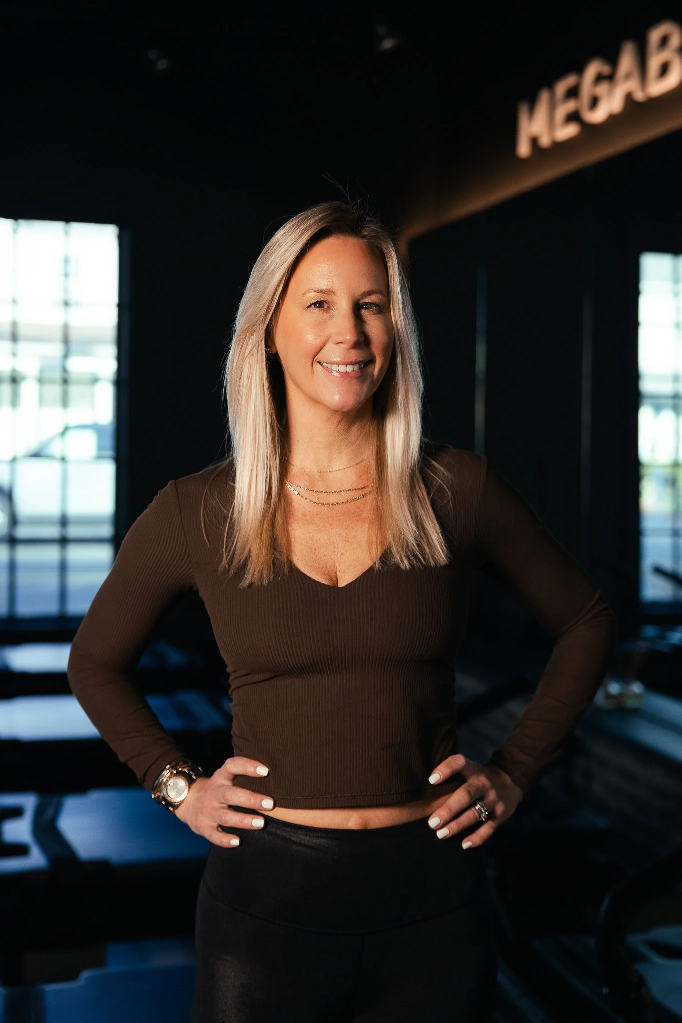 A woman with long blonde hair standing in a dark room with large windows and a sign reading 'MEGAB'. She is wearing a brown long-sleeve top, black skirt, and jewelry, smiling with hands on hips.