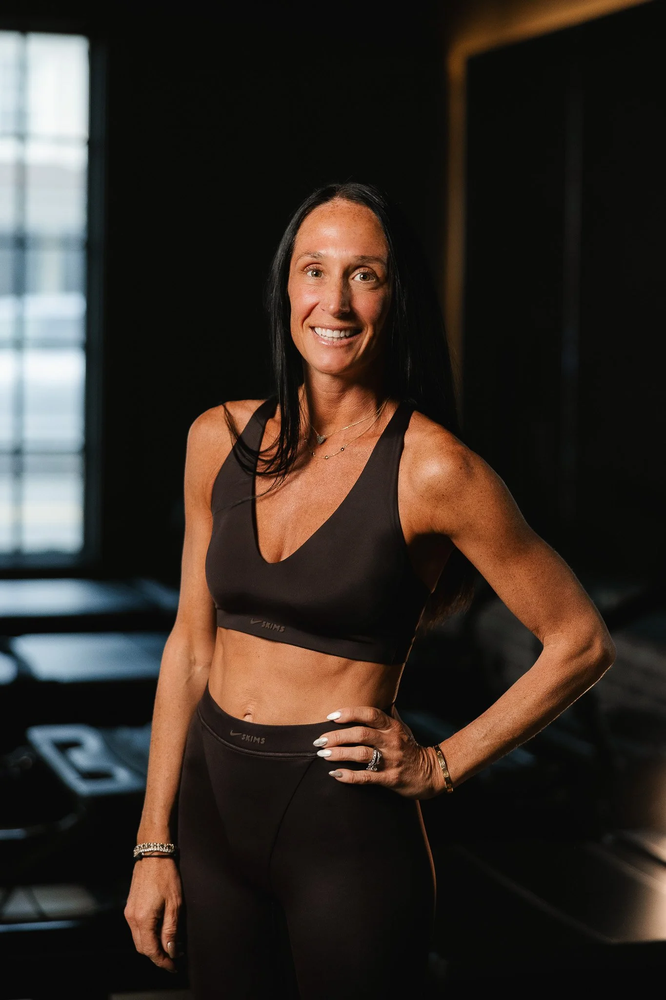 A woman in athletic wear smiling inside a gym, with workout equipment in the background.