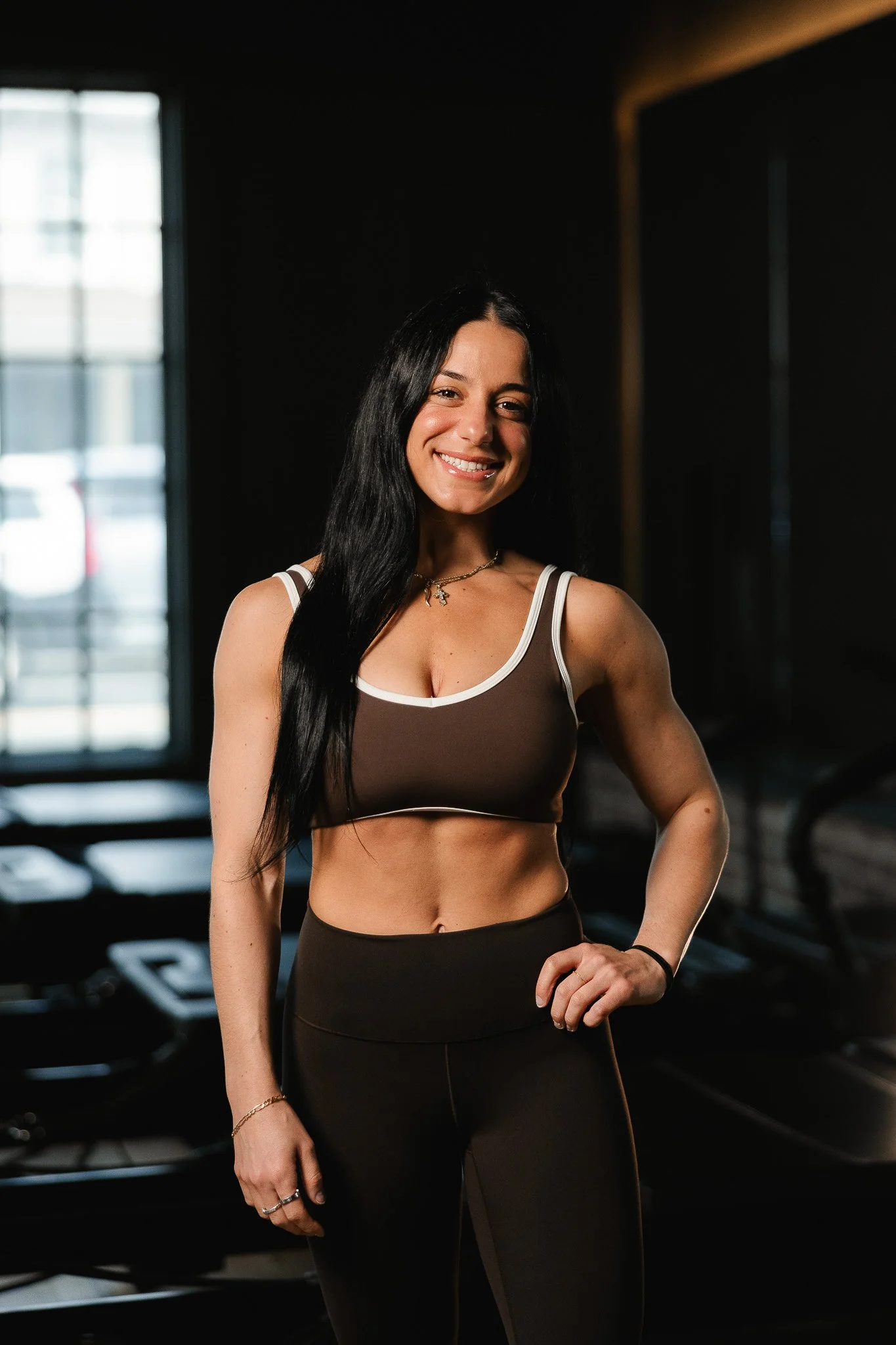 A woman with long black hair in a gym, smiling and wearing a brown sports bra with white straps and brown workout leggings.