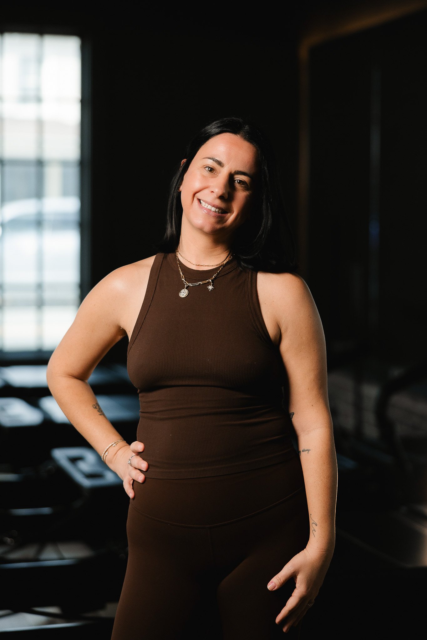 A woman with black hair, wearing a brown sleeveless top and jewelry, smiling with her hand on her waist inside a dimly lit gym.