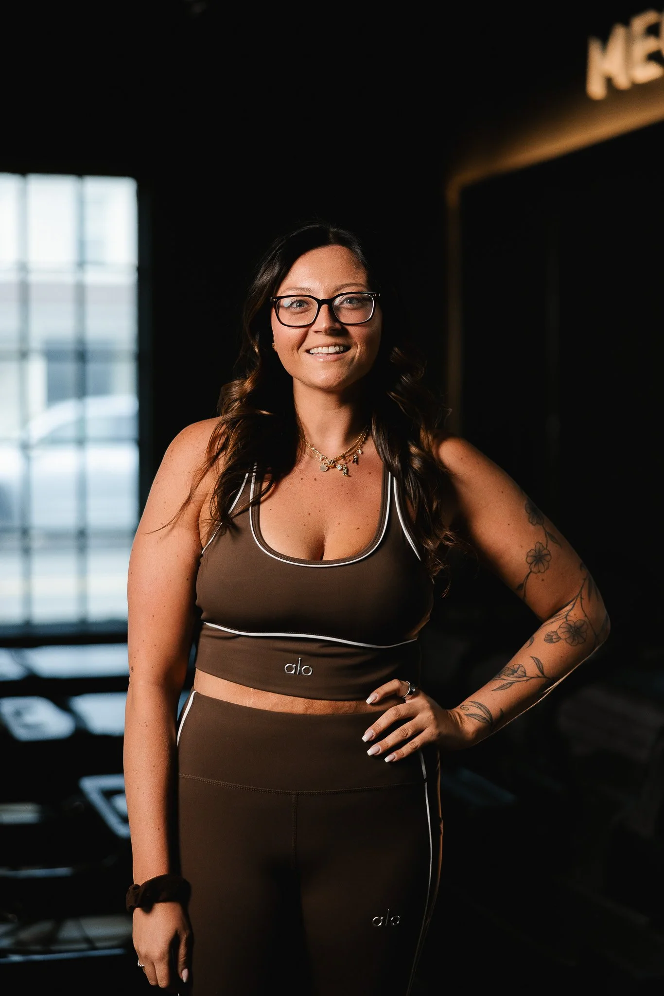 A woman with long dark hair, glasses, and tattoos on her arm and side, standing in a gym wearing a brown sports bra and matching leggings with white details, smiling at the camera.