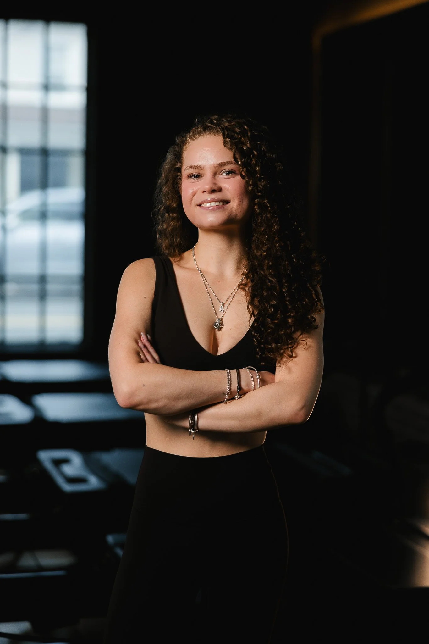 Portrait of a smiling woman with curly hair, wearing a black sleeveless top and jewelry, standing in a dark indoor gym with windows in the background.