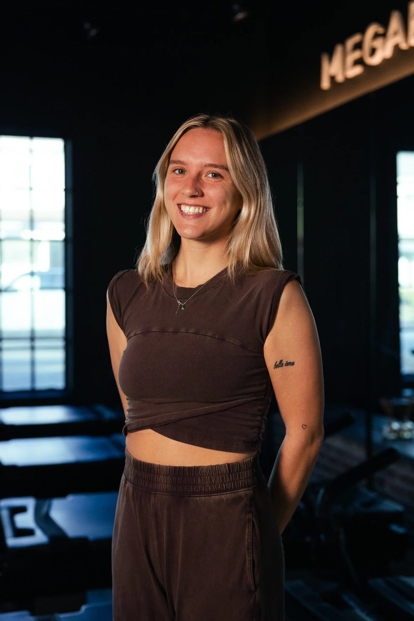 A young woman with blonde hair and a nose ring smiling in a gym. She is wearing a brown athletic crop top and brown pants, with tattoos on her left arm. The gym has large windows and gym equipment in the background.