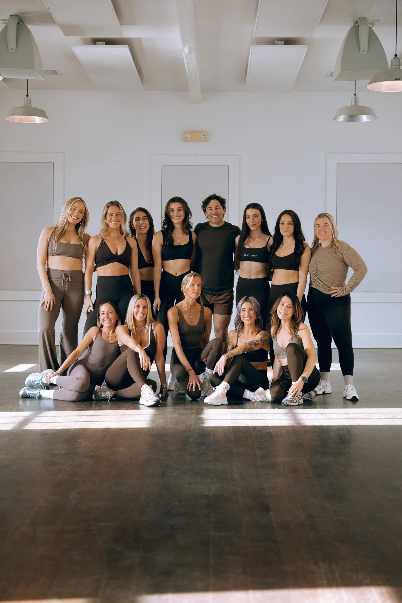 A group of women and one man posing for a photo in a dance studio with hardwood floors and white walls. The women are dressed in athletic workout clothes, mostly in black or neutral tones, with some kneeling or sitting in the front row and others standing behind. The man is standing in the middle, smiling. There is sunlight coming through the window, creating stripes on the floor.