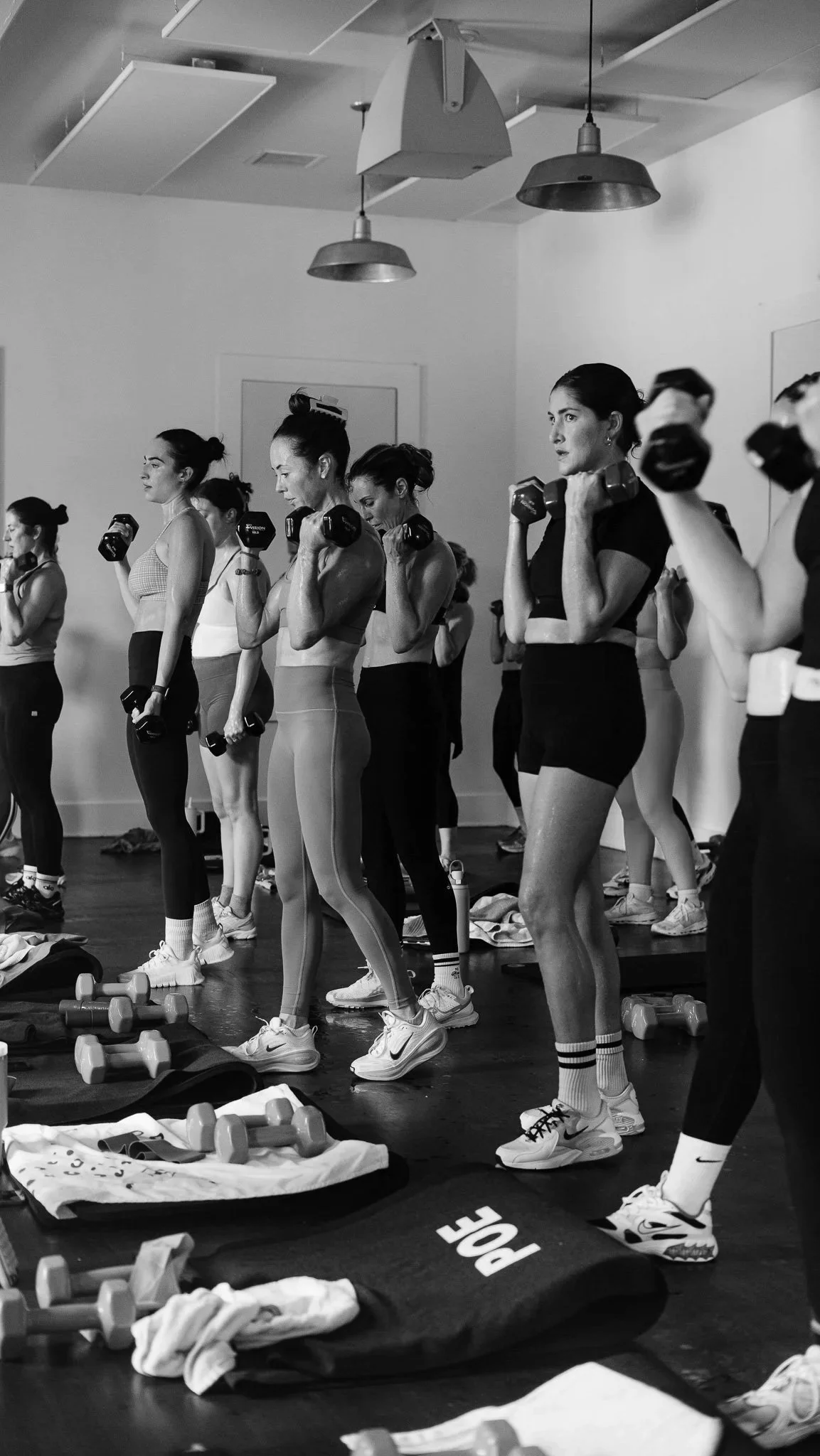 Women participating in a group fitness class lifting dumbbells in a studio, with workout gear and water bottles on the floor.