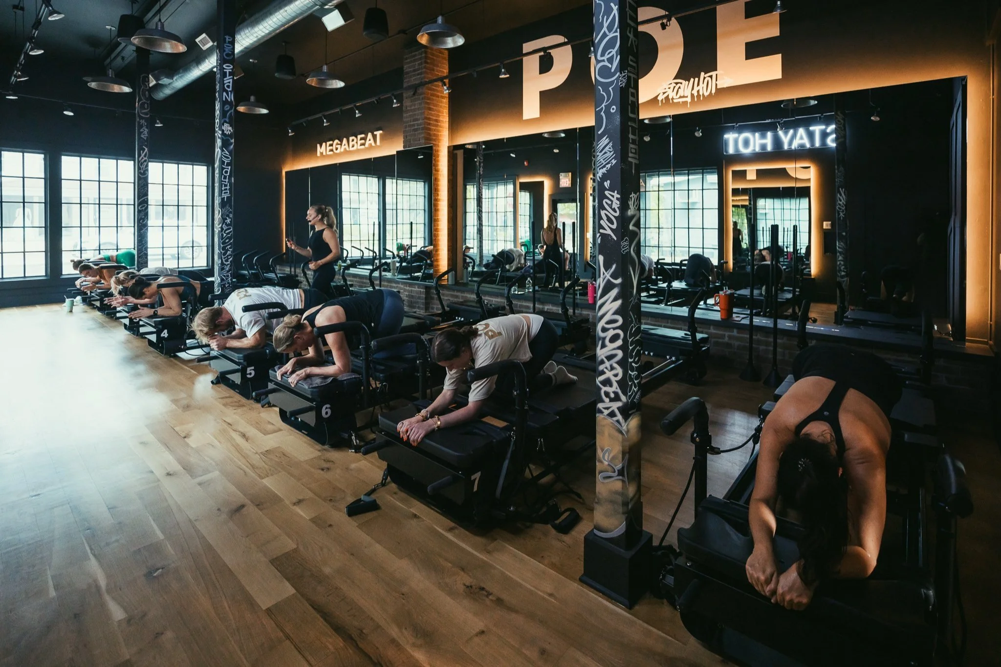 People in a fitness class doing planks on exercise benches inside a gym with large windows and industrial decor.
