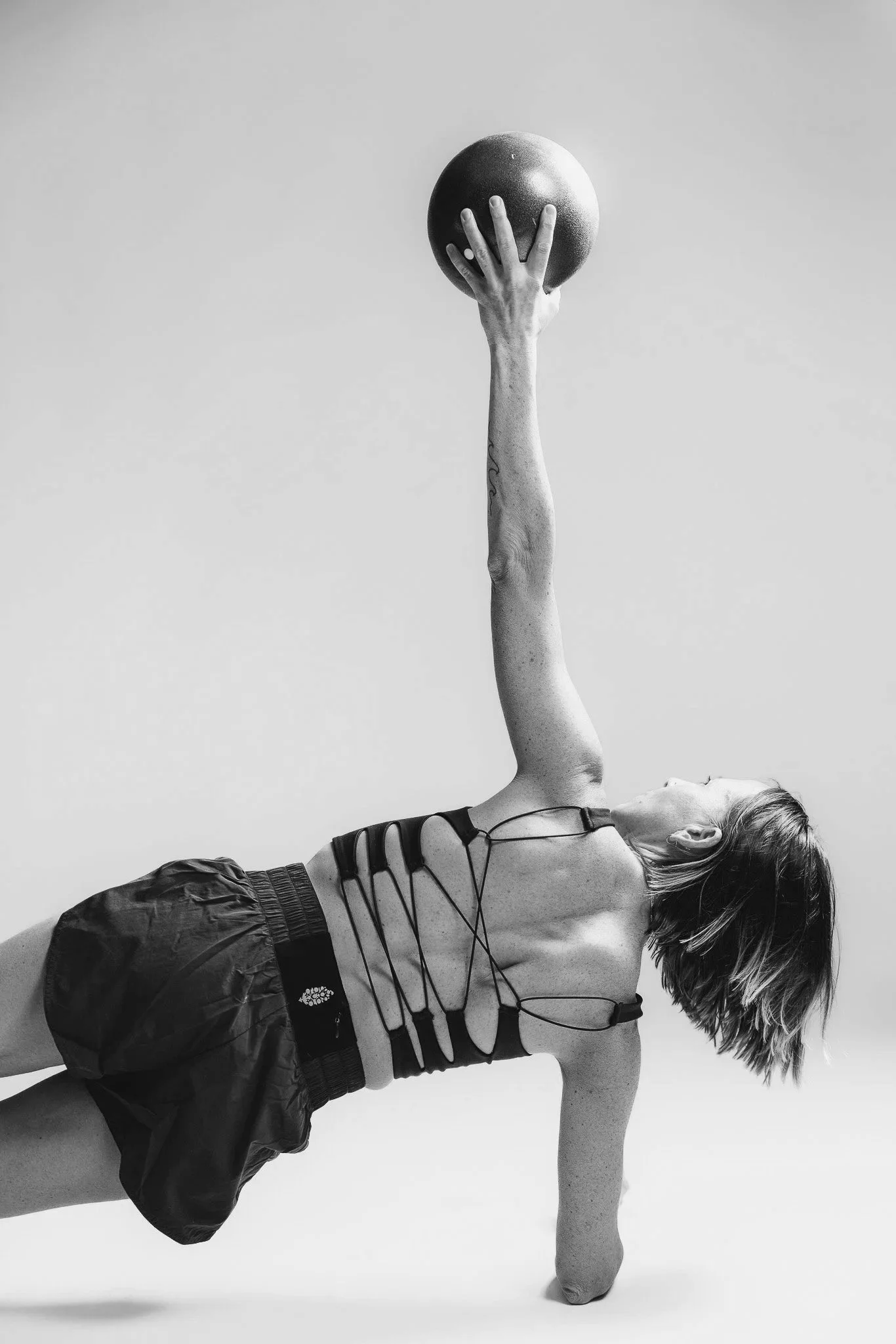 A person with light skin and short hair is lying on their side, supporting themselves with one arm and holding a basketball high above their head with their other hand. The person is wearing a sports bra and shorts, with a tattoo visible on their inner forearm. The background is plain and the image is in black and white.