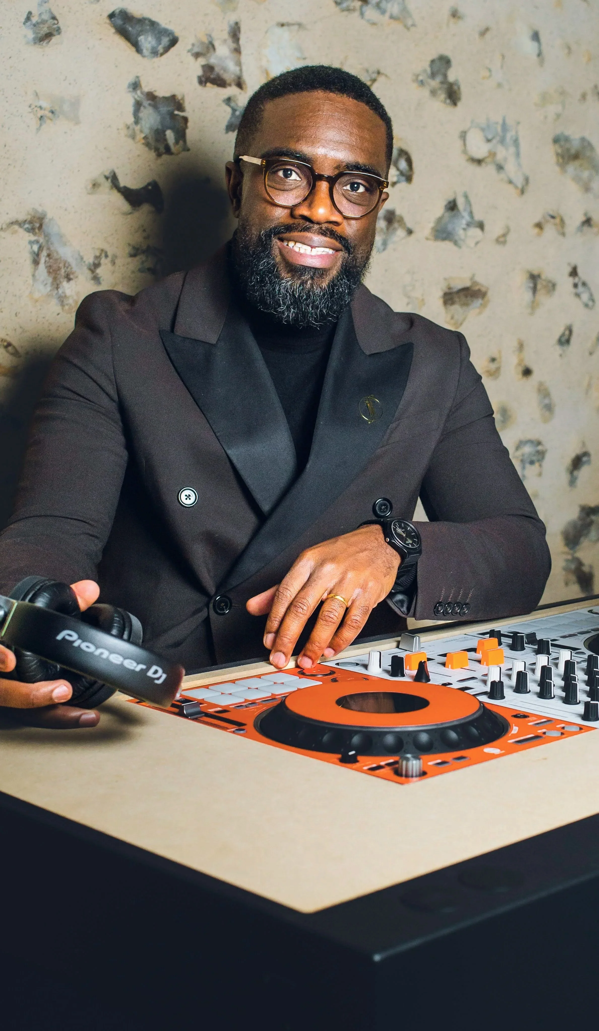 Un homme noir portant des lunettes, un costume sombre, et un bracelet montre, pose derrière un éclairage DJ Pioneer au centre d'une table, en train de mixer ou de préparer une musique.
