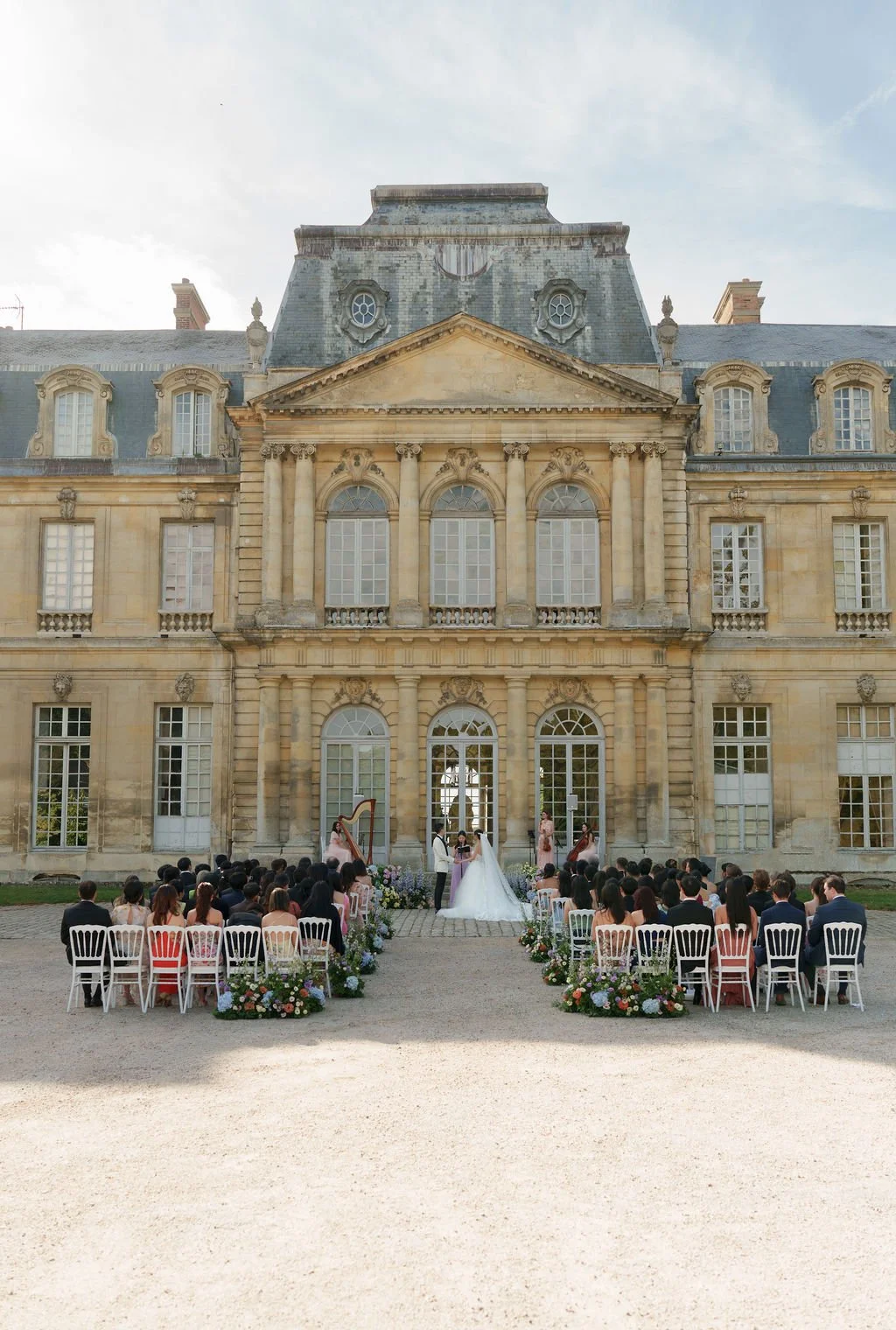 Cérémonie de mariage en plein air devant un grand bâtiment historique avec des invités assis, une mariée et un marié qui s'échangent leurs vœux, entourés de fleurs.