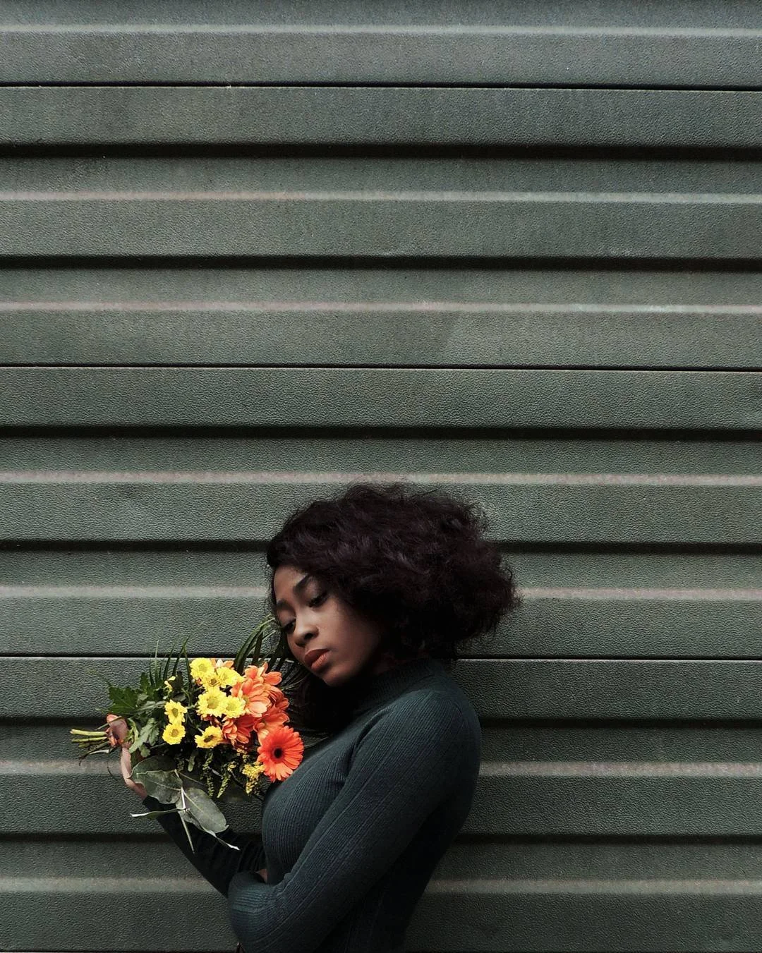 Une femme avec des cheveux bouclés bruns foncés, portant un haut noir, tient un bouquet de fleurs colorées dont des œillets, des marguerites et des roses, devant un mur en métal à stries horizontales.