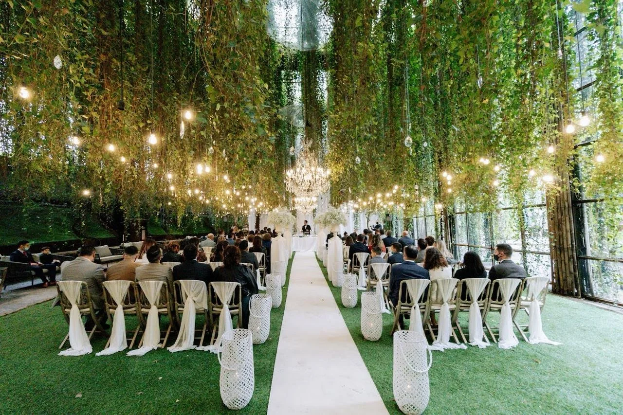 Cérémonie de mariage dans un espace vert intérieur décoré avec des guirlandes lumineuses et des végétaux suspendus, avec des invités assis face à l'autel