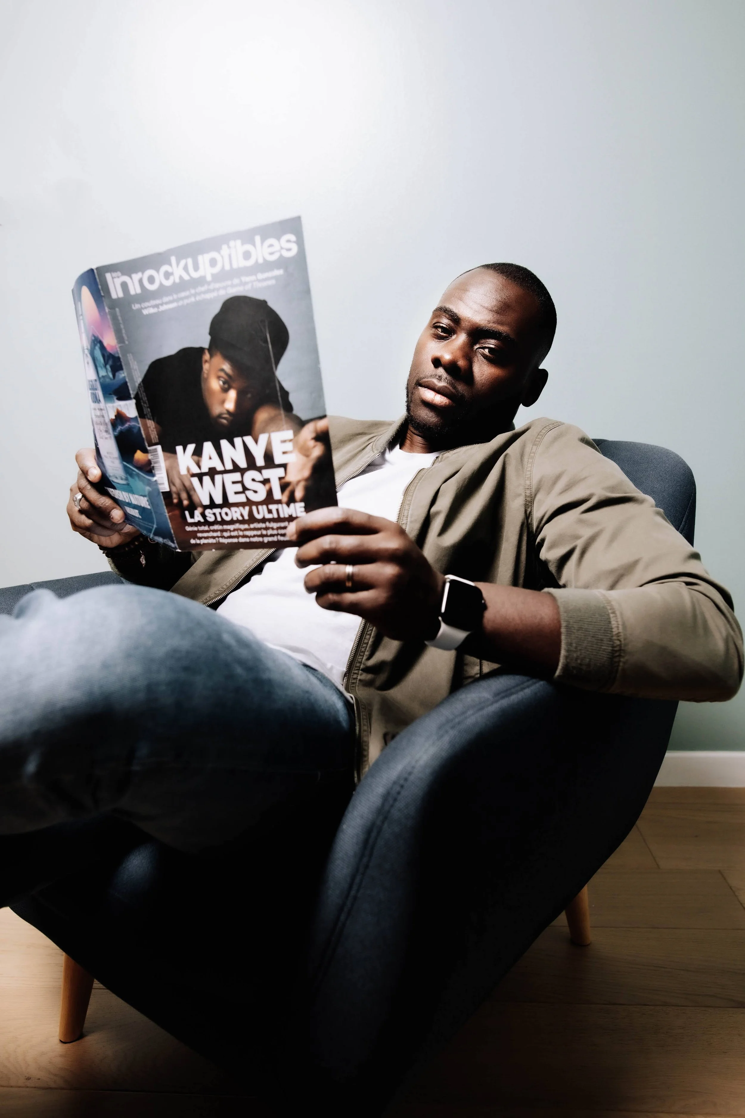 Un homme assis dans un fauteuil noir, portant une veste beige, une t-shirt blanc, et une montre connectée, tient un magazine avec une photo de Kanye West sur la couverture.