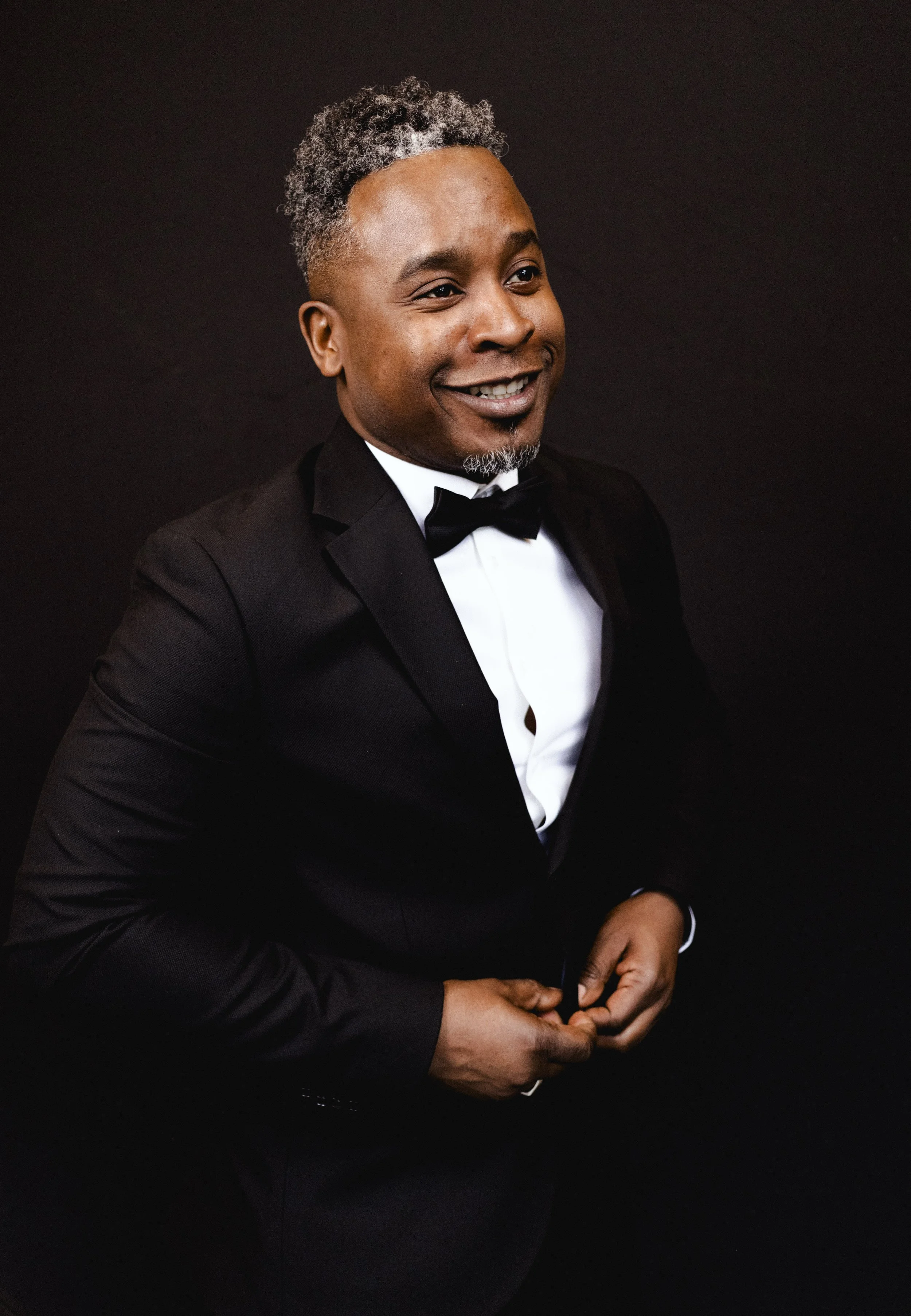 Homme souriant en smoking avec nœud papillon, posant contre un fond noir.