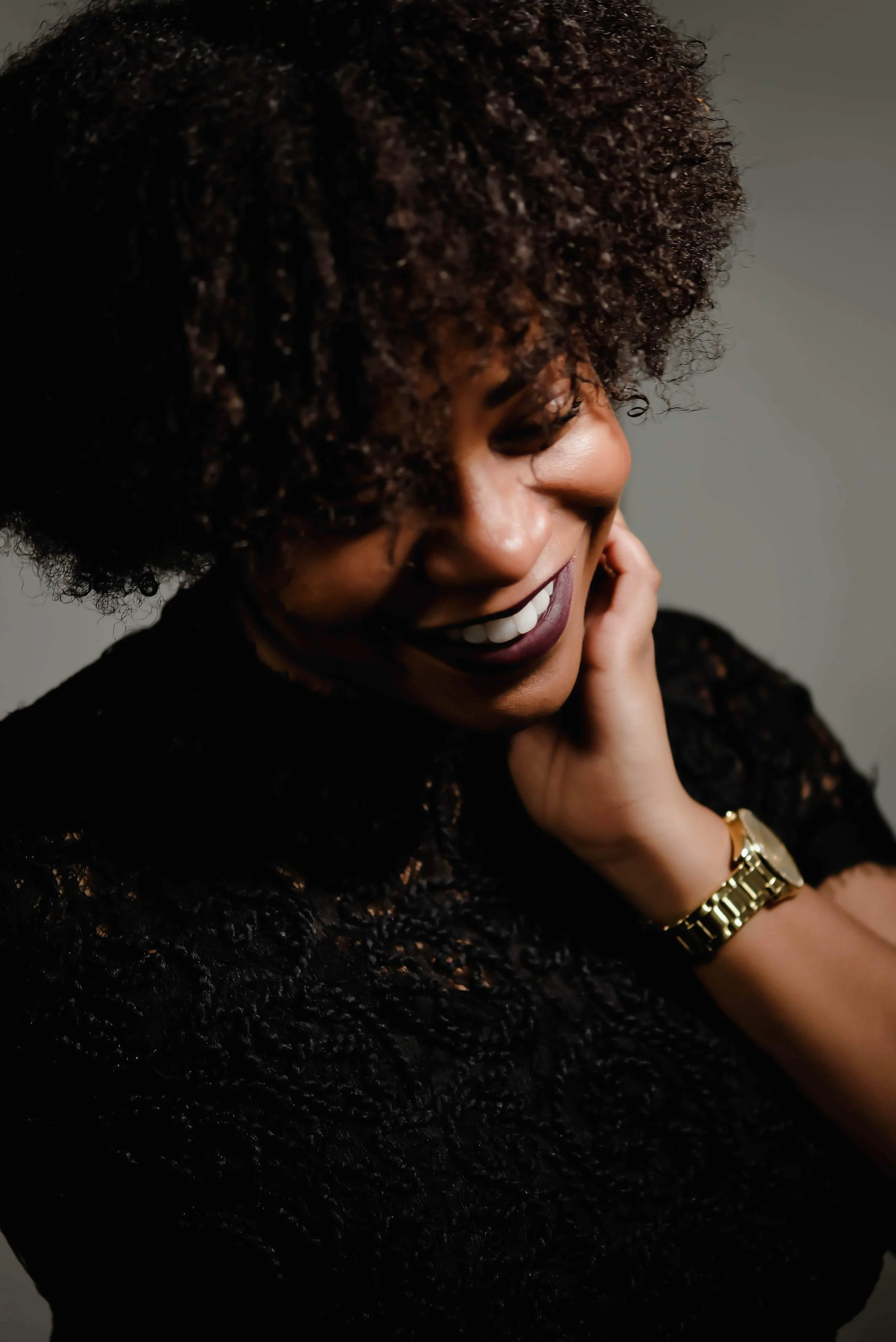 Jeune femme avec des cheveux bouclés, souriant, portant un maquillage foncé et une montre dorée, vêtue d'une robe en dentelle noire.