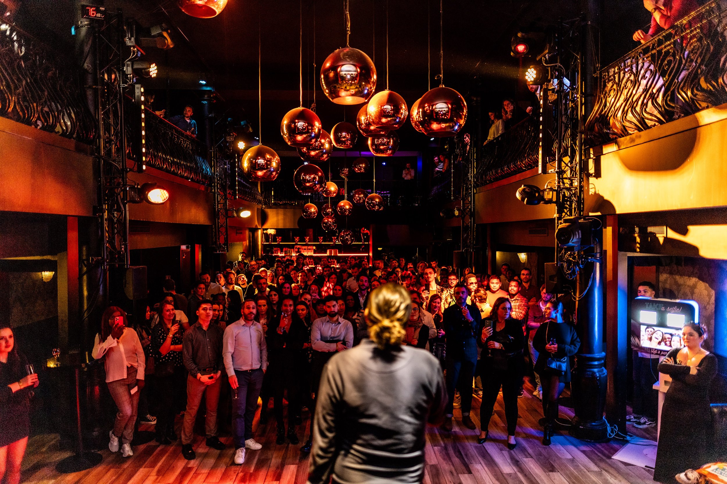 Une scène de discothèque animée avec un DJ ou un animateur en avant-plan, un grand public de personnes assises et debout, décorations avec des ballons dorés suspendus au plafond, éclairage tamisé, et un espace de bar en arrière-plan.