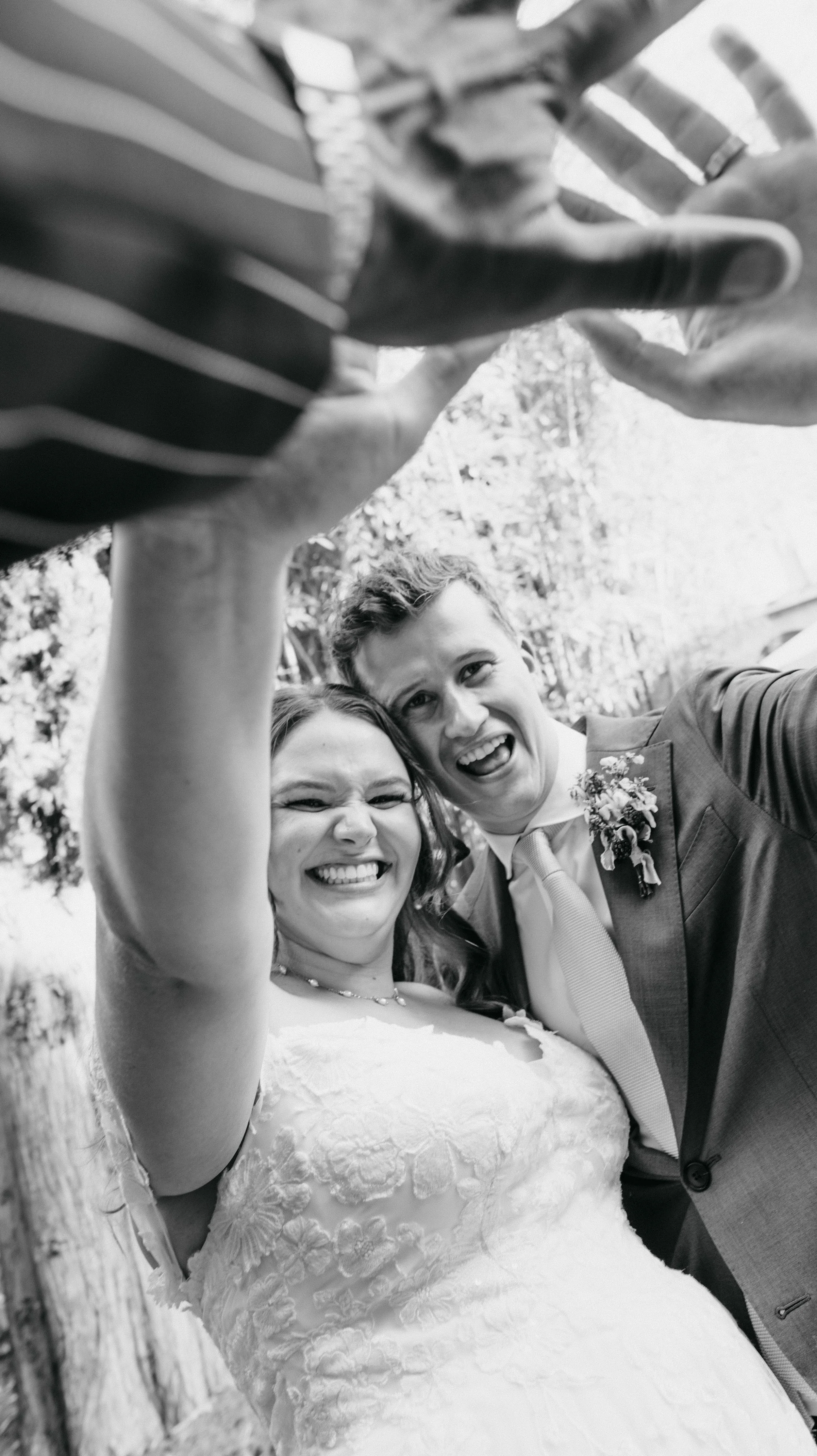 Photo en noir et blanc d'un couple de mariage souriant et prenant un selfie ensemble, la femme portant une robe de mariée et l'homme en costume avec une boutonnière.