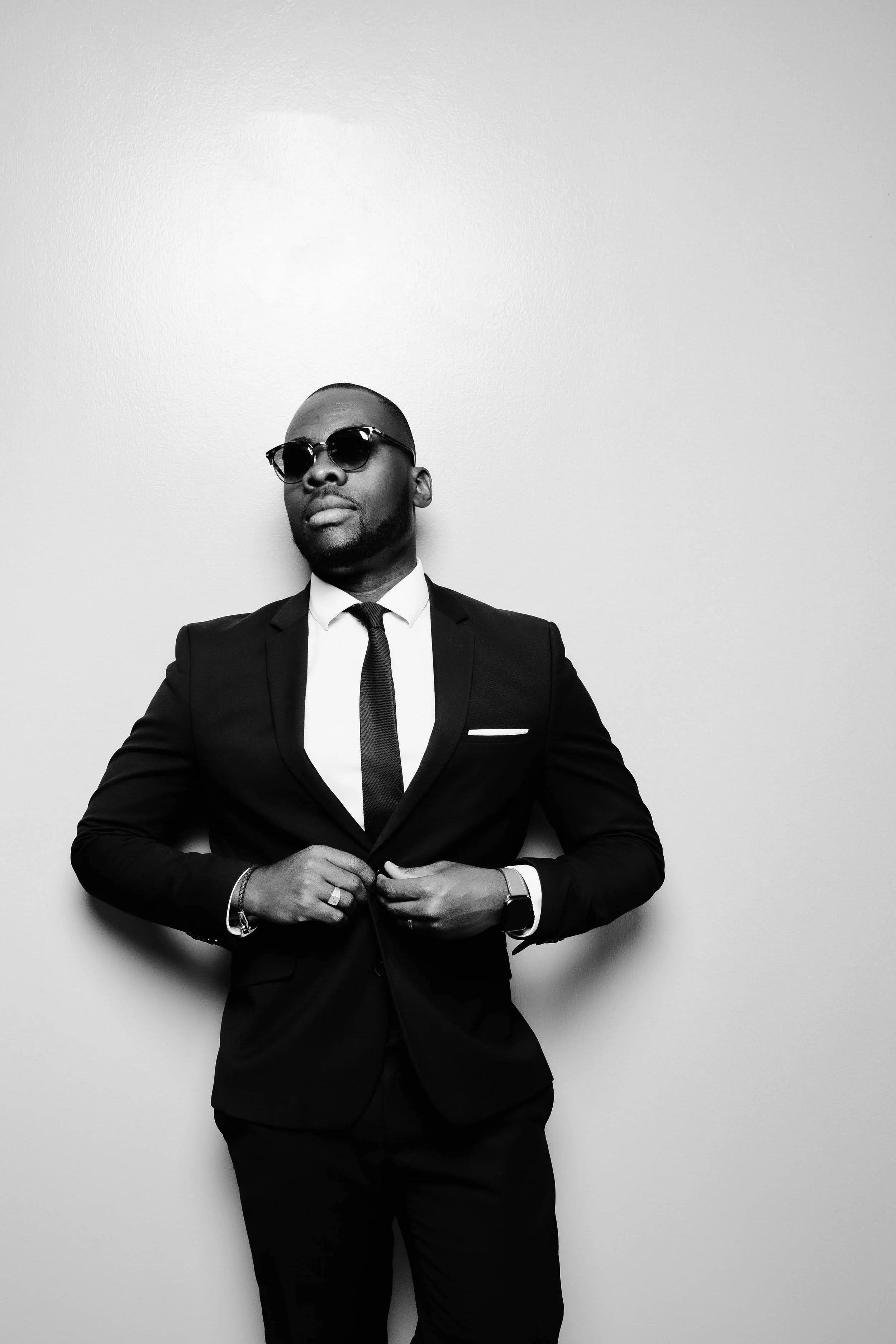 Homme en costume noir, lunettes de soleil, pose confiante contre un mur blanc