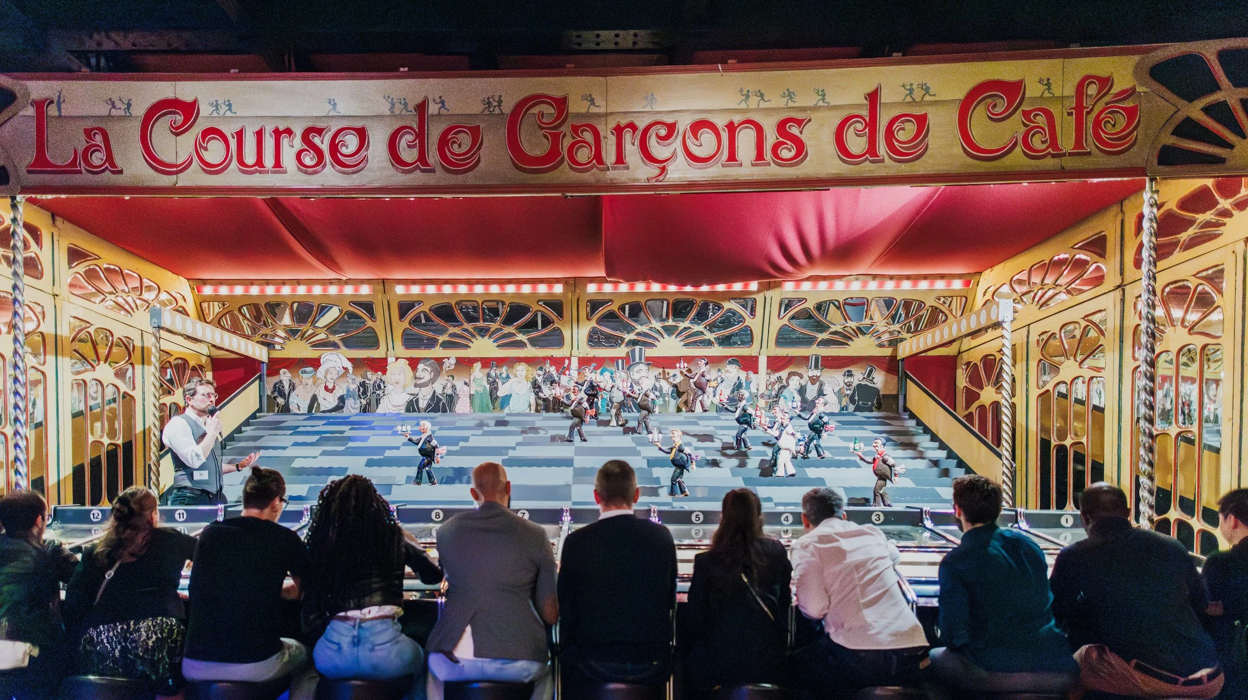 Une scène de spectacle de marionnettes représentant une fête ou un carnaval, avec des marionnettes habillées en costumes, derrière un décor coloré et animé, sous une tente rouge avec le panneau 'La Course de Garçons de Café'.