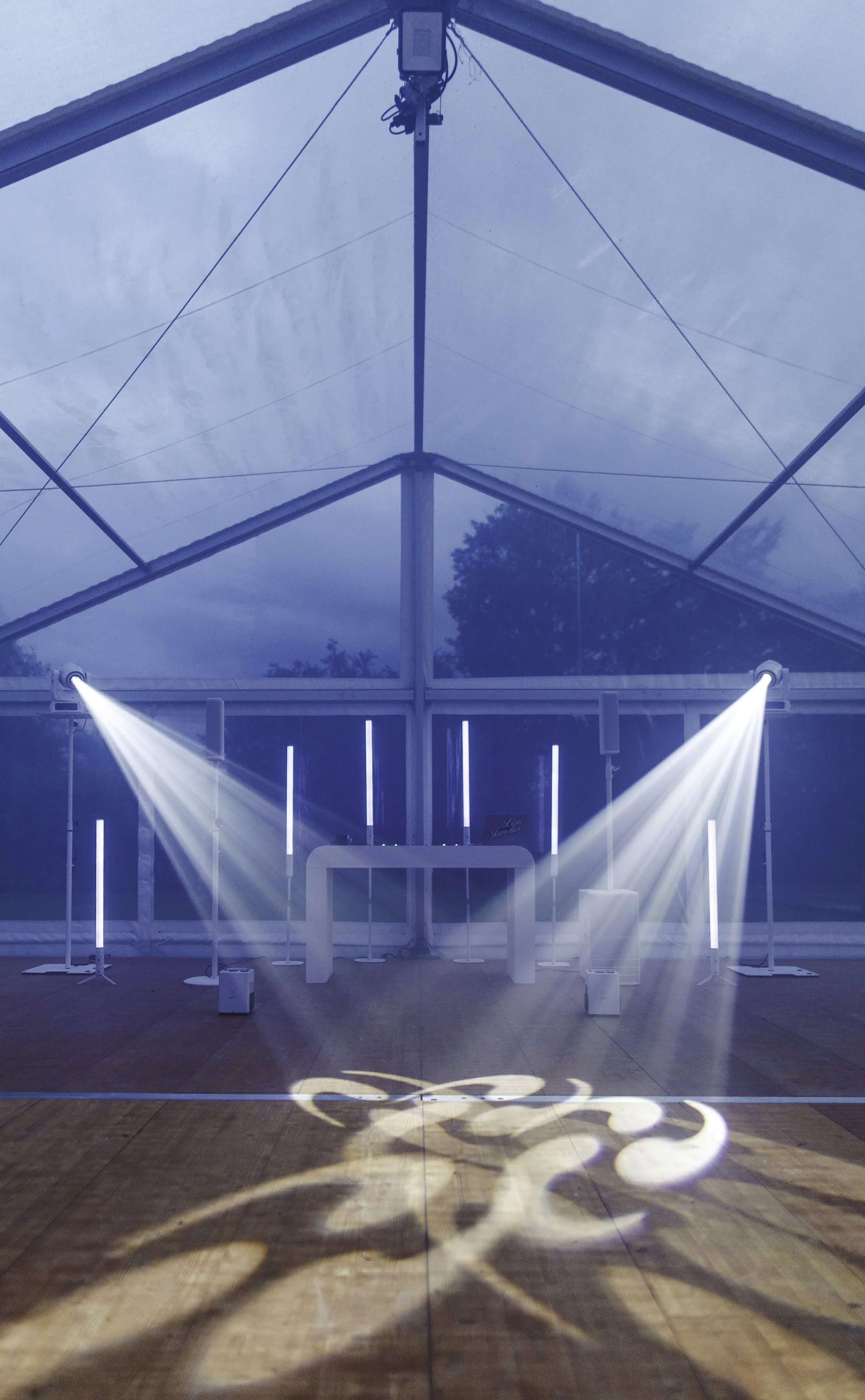 Intérieur d'une tente ou d'une structure en plein air avec éclairage de scène, plancher en bois et écran au fond, avec des lumières de scène et des rayons lumineux projetés au sol.