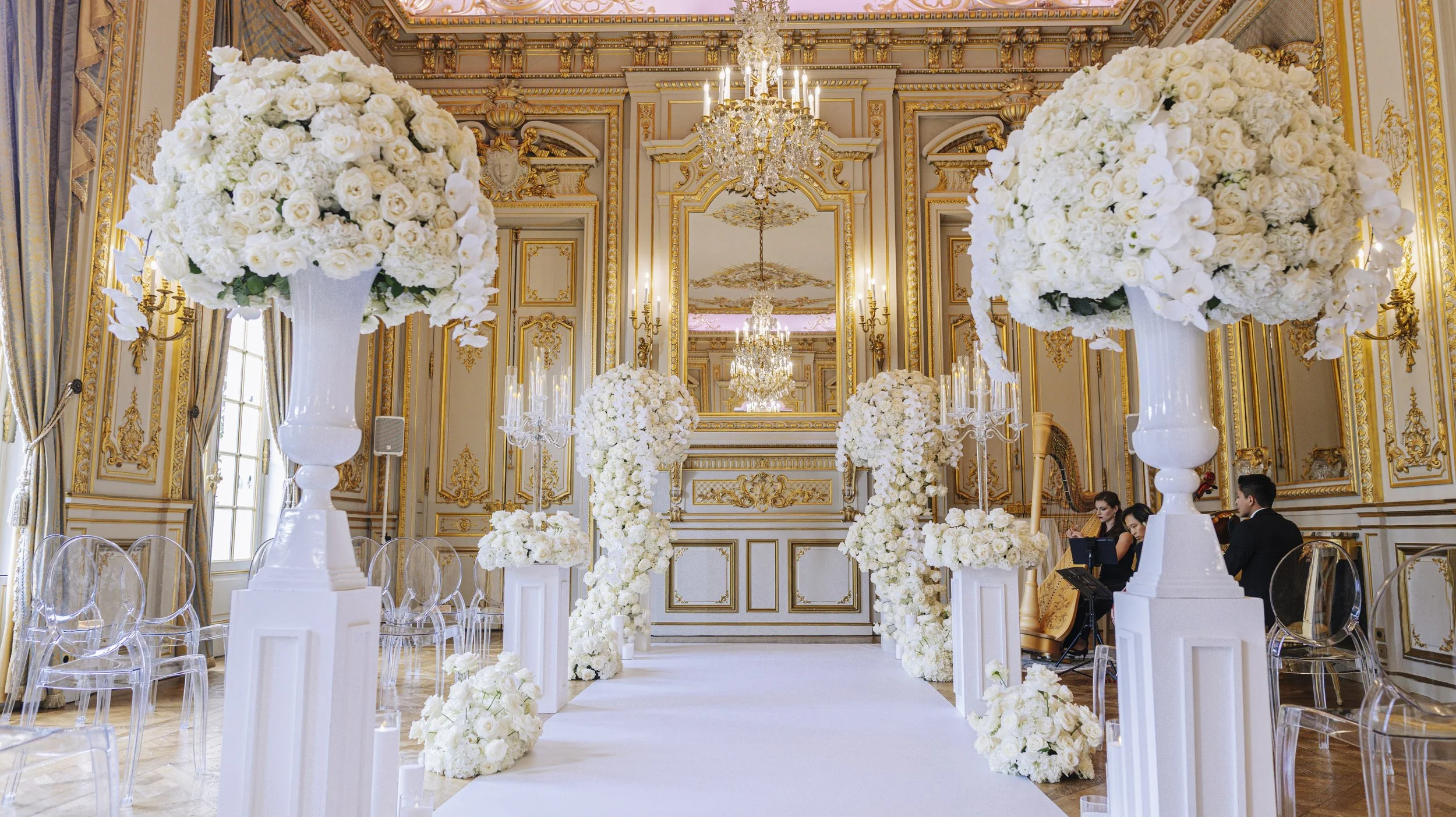 Salle de mariage élégante avec décorations florales blanches, chandeliers en cristal, grands miroirs et ornements dorés. Chemin de fleurs blanches et chaises en acrylique transparent. Musiciens en arrière-plan.