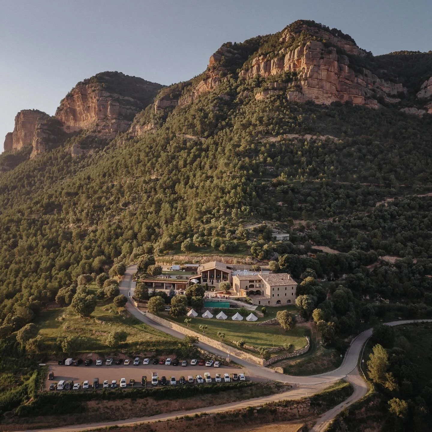 Vue aérienne d'une maison de luxe située dans une région montagneuse entourée d'arbres, avec un parking rempli de voitures et une piscine à proximité.