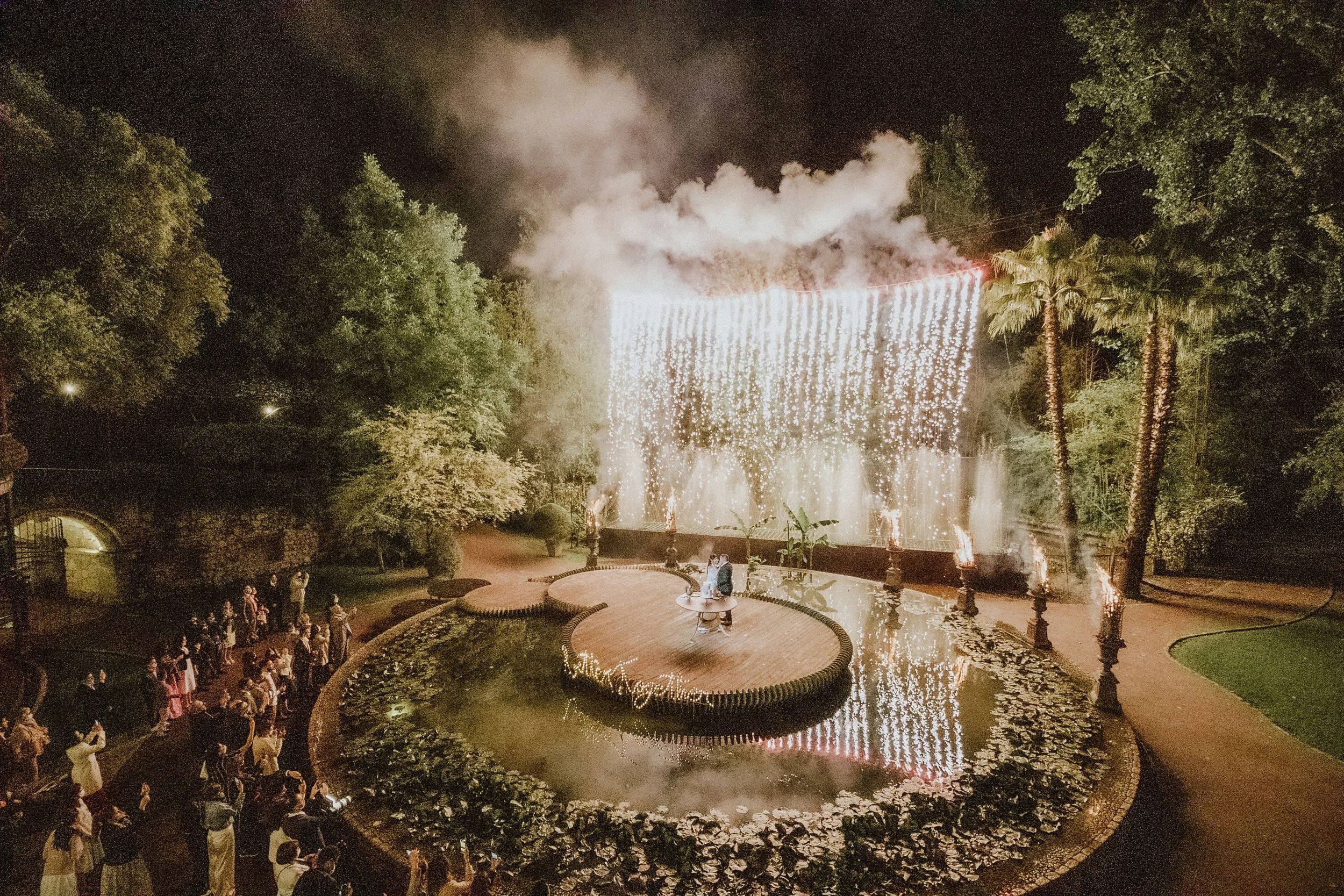 Un mariage avec un feu d'artifice dans un parc la nuit, où deux personnes sont assises sur une petite plateforme ronde entourée d'une fontaine et de fleurs, avec une foule de spectateurs autour.