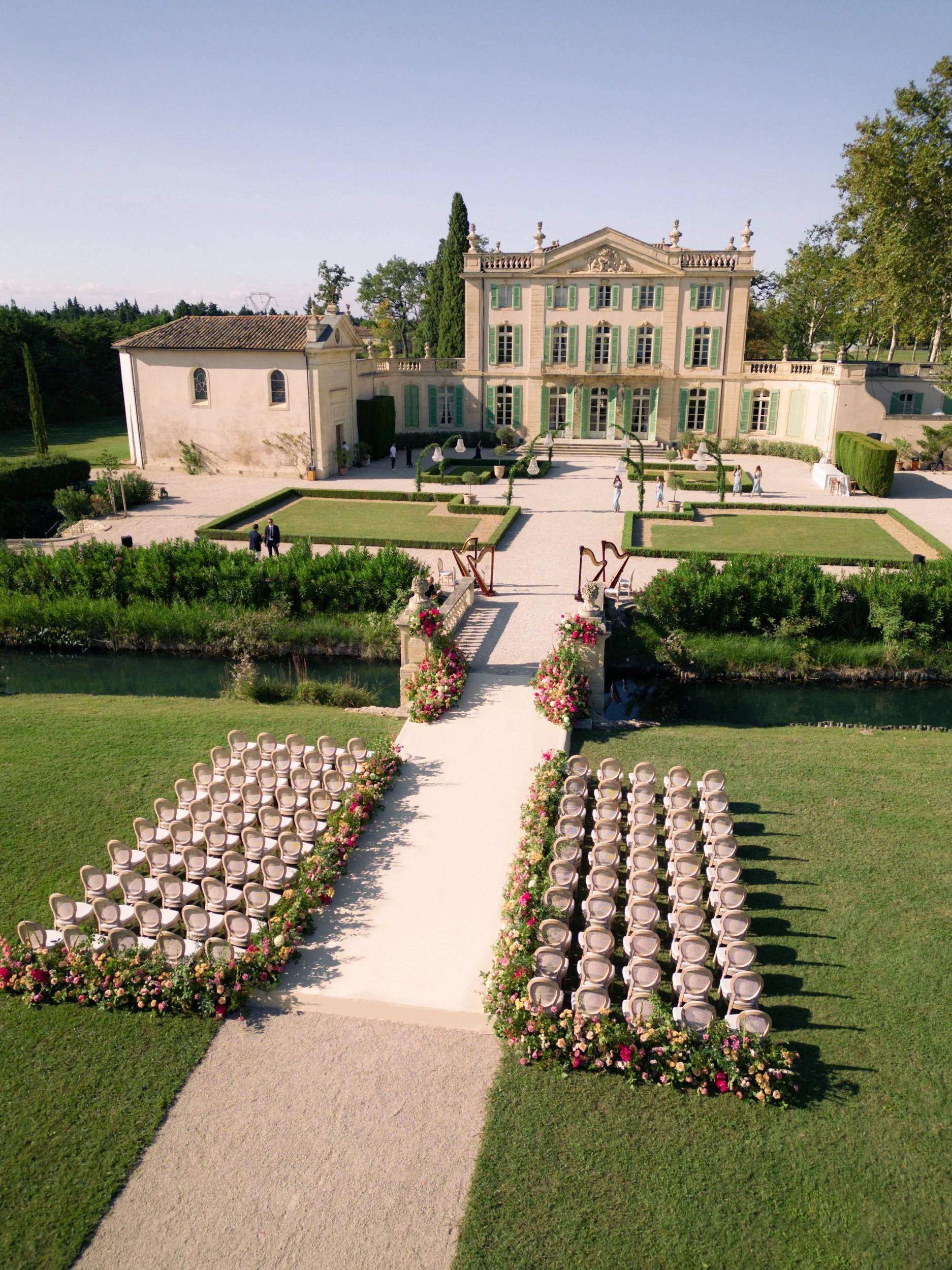 Un endroit extérieur décoré pour un mariage, avec des chaises disposées de part et d'autre d'une allée centrale bordée de fleurs colorées, menant à un grand bâtiment élégant avec des fenêtres à volets verts et un jardin bien entretenu.