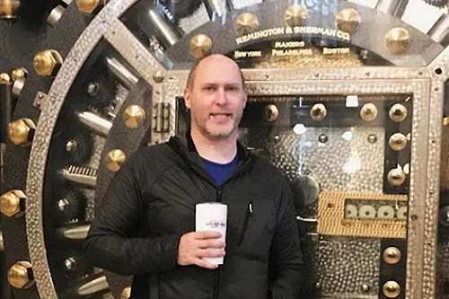 Tim Gill, SEO and Marketing expert, holding a drink standing in front of a large, circular vault door with gold and silver details.
