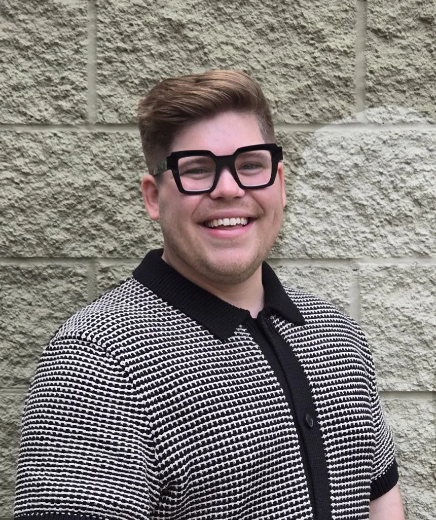 A young man with light skin, wearing large black glasses, smiling, and standing in front of a light-colored textured brick wall.